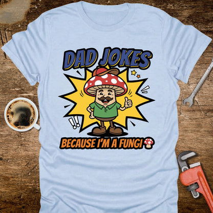 Light blue t-shirt with a cartoon mushroom character and text on a wooden surface with a coffee cup and wrench.
