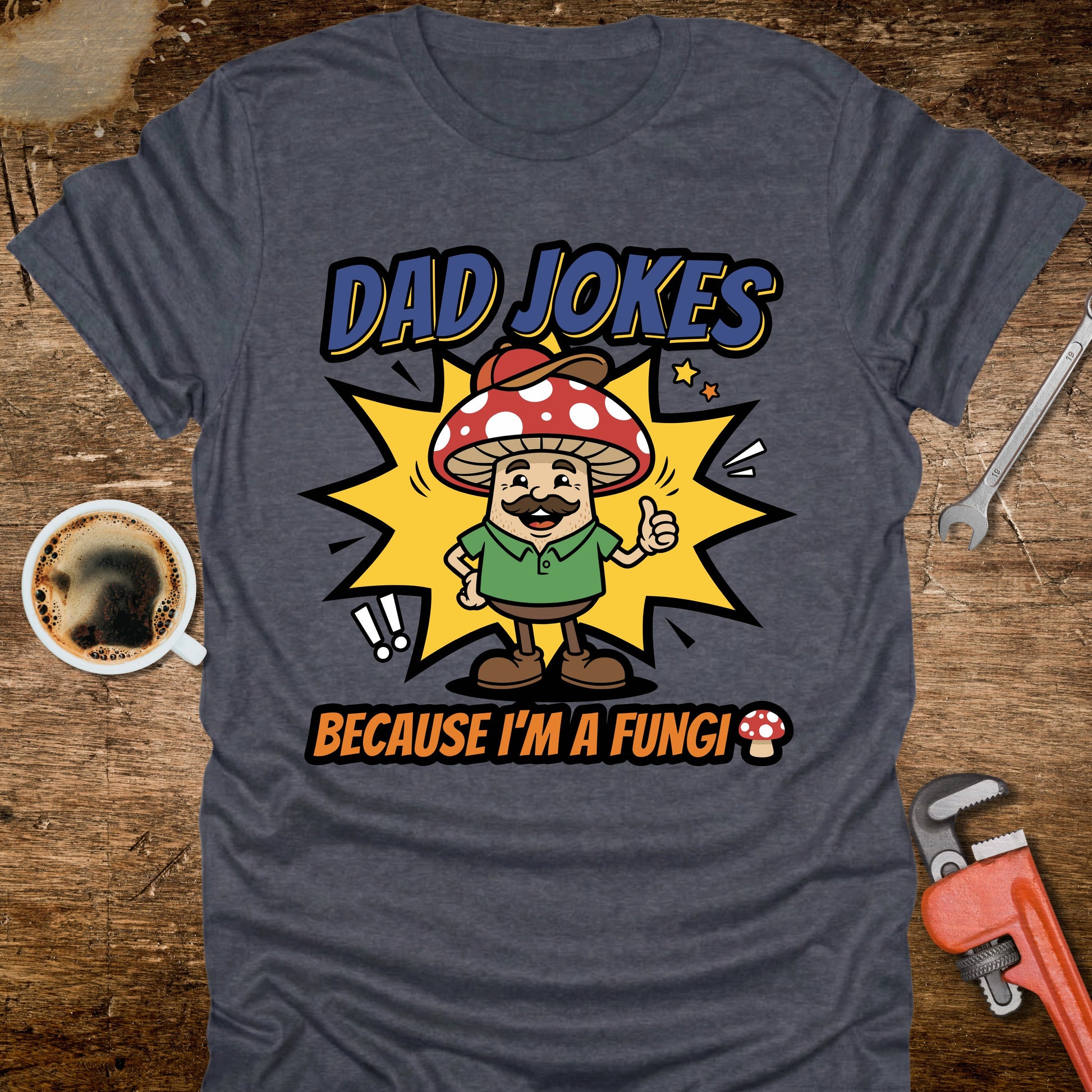Gray t-shirt with a mushroom graphic and text 'Dad jokes because I'm a fungi' on a wooden surface with coffee and tools.