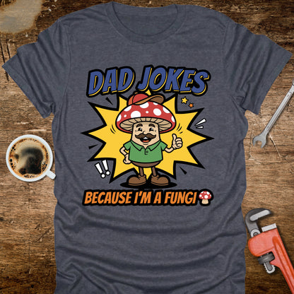 Gray t-shirt with a mushroom graphic and text 'Dad jokes because I'm a fungi' on a wooden surface with coffee and tools.