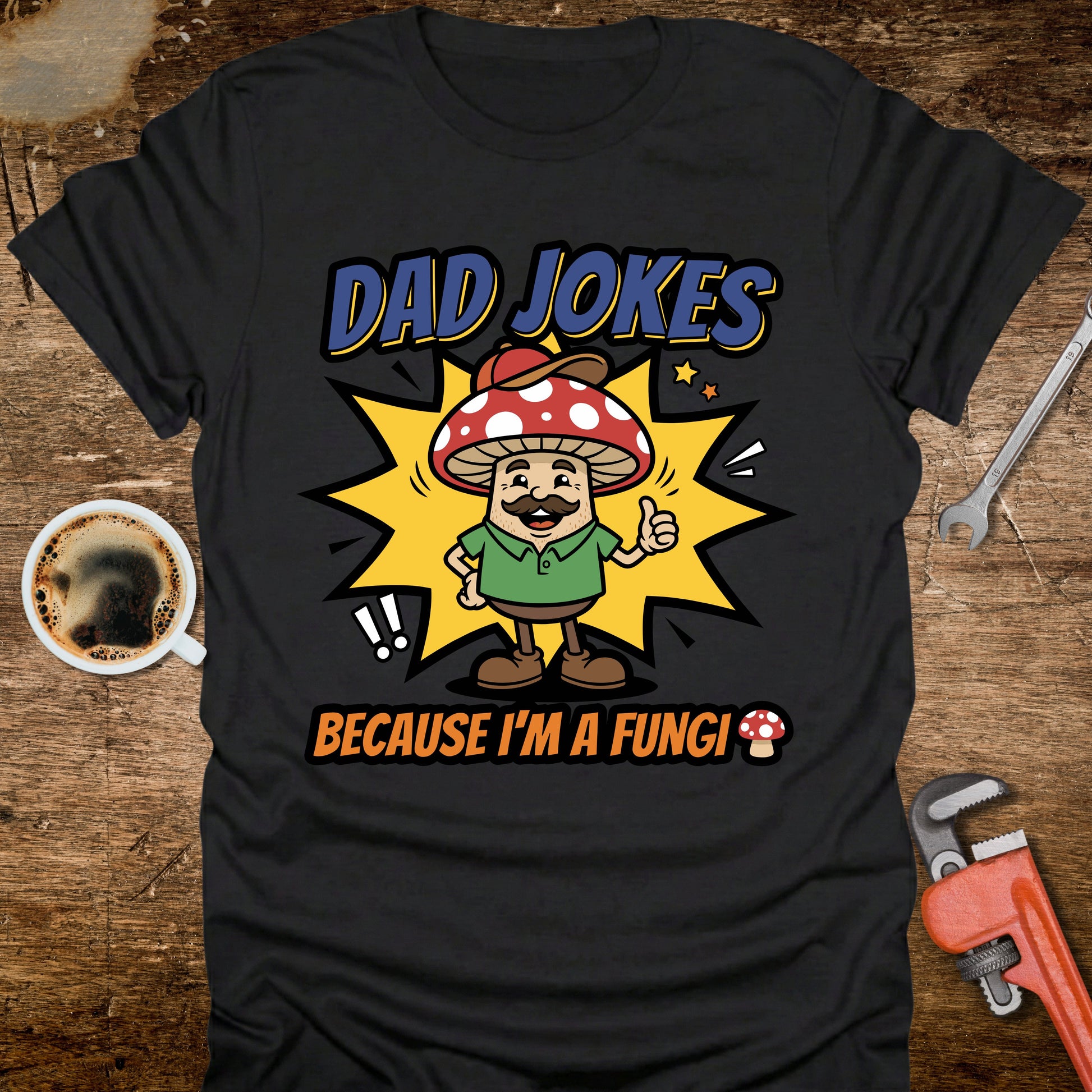 Black t-shirt with a cartoon mushroom character and text 'Dad jokes because I'm a fungi' on a wooden surface with tools and coffee.