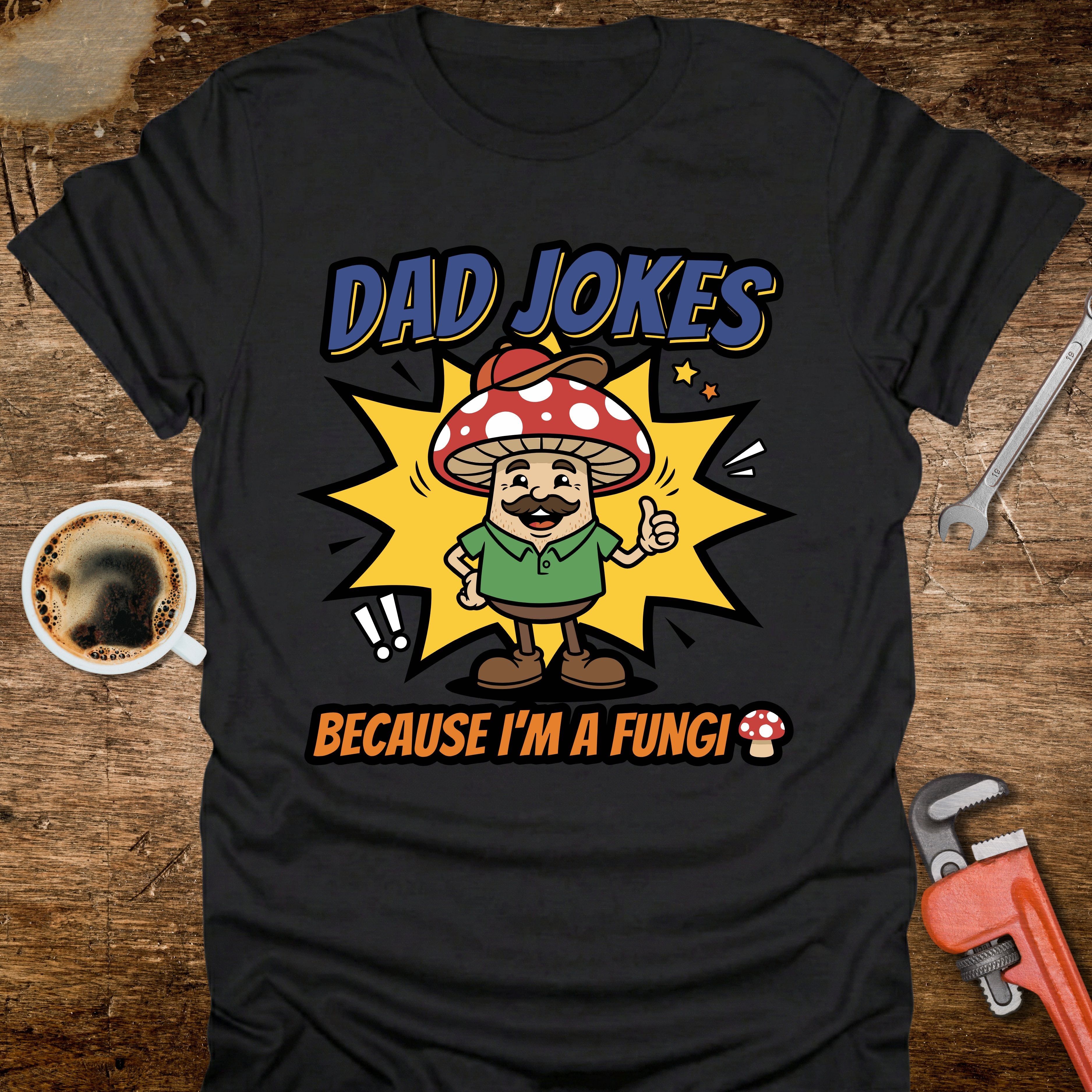 Black t-shirt with a cartoon mushroom character and text 'Dad jokes because I'm a fungi' on a wooden surface with tools and coffee.
