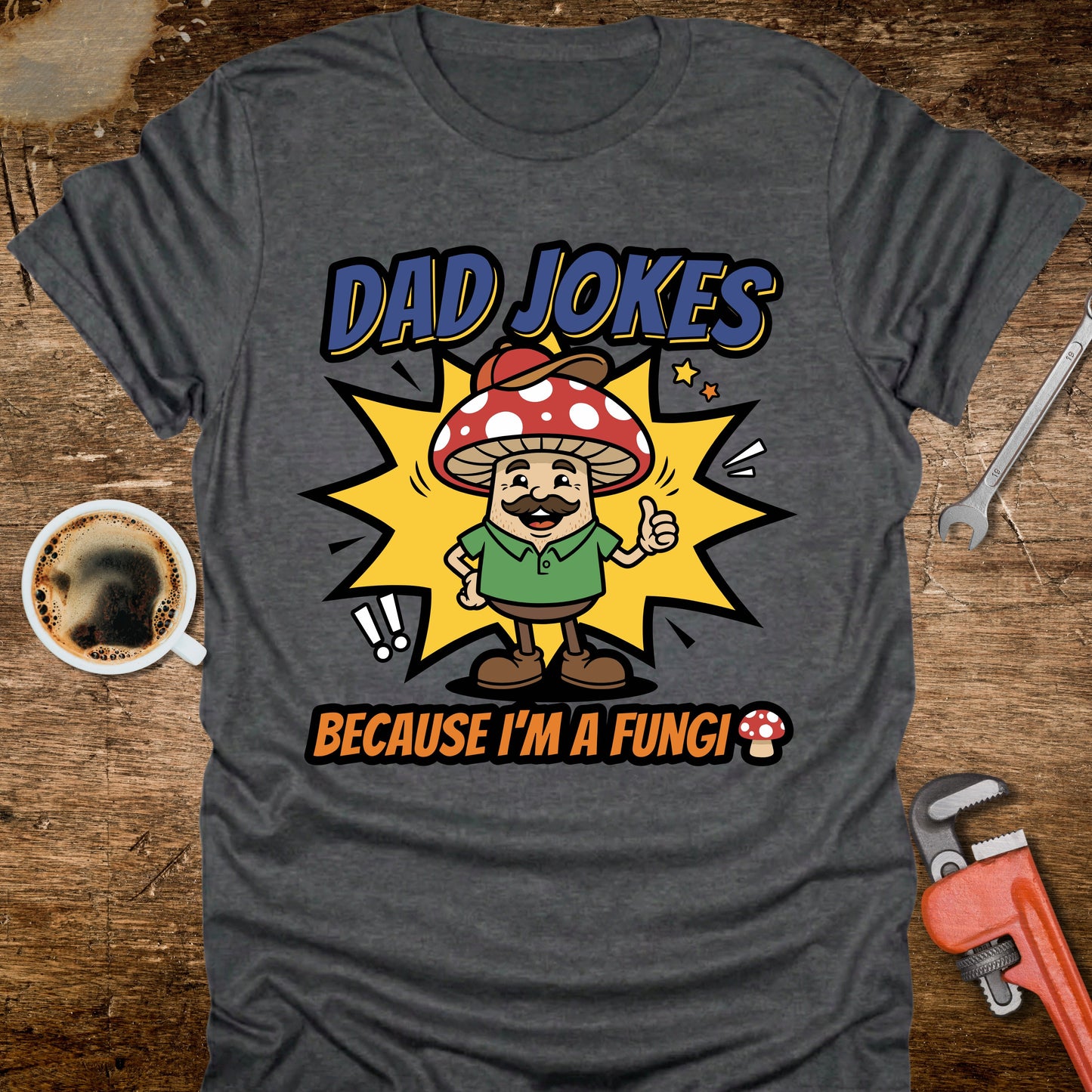Gray t-shirt with a mushroom graphic and text 'Dad Jokes Because I'm a Fungi' on a wooden surface with coffee and tools.