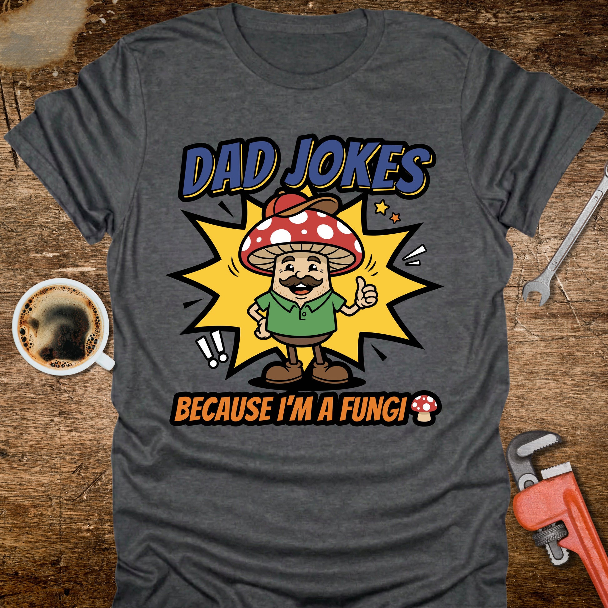 Gray t-shirt with a mushroom graphic and text 'Dad Jokes Because I'm a Fungi' on a wooden surface with coffee and tools.