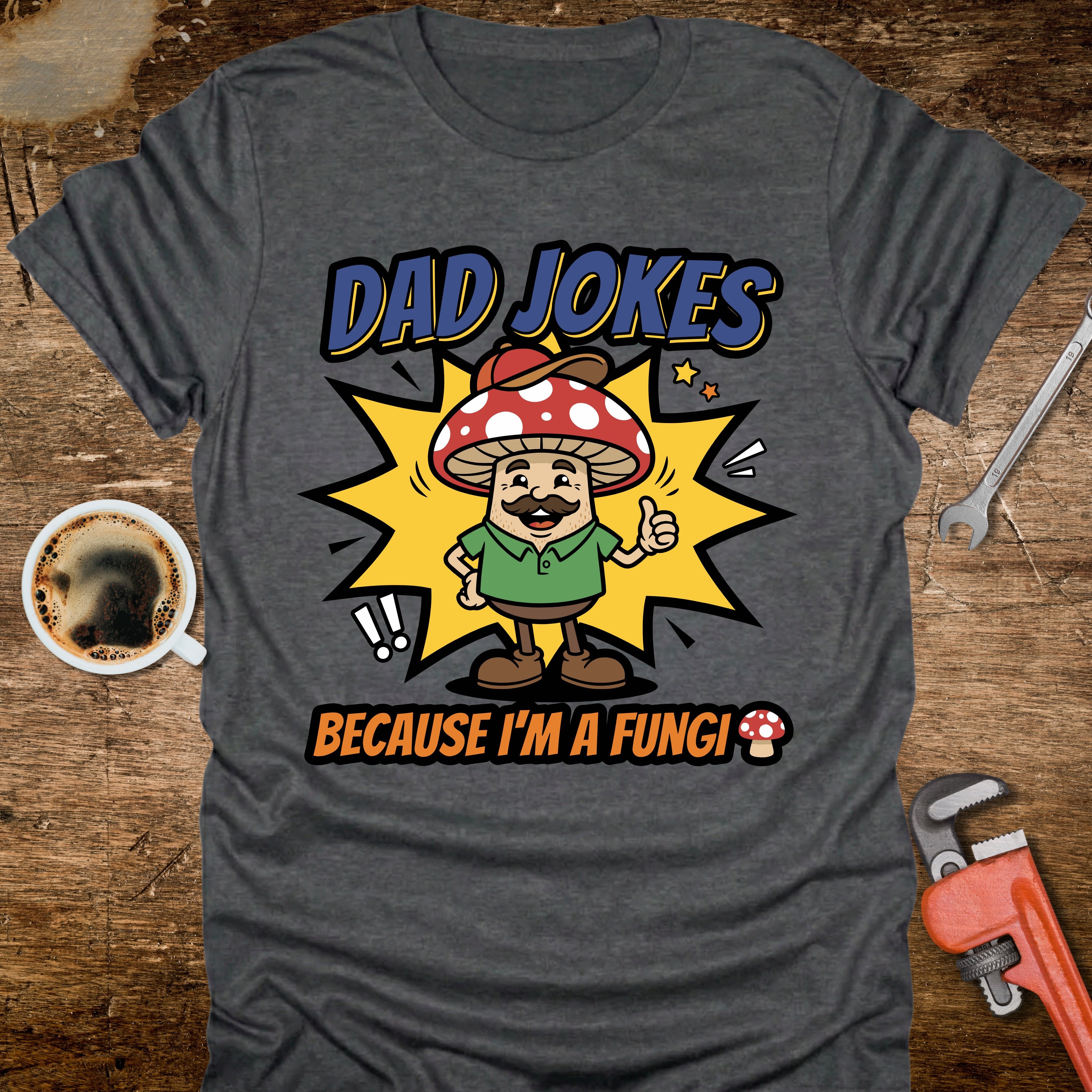 Gray t-shirt with a mushroom graphic and text 'Dad Jokes Because I'm a Fungi' on a wooden surface with coffee and tools.