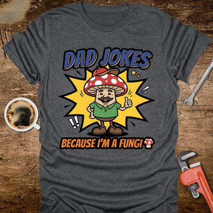 Gray t-shirt with a mushroom graphic and text 'Dad Jokes Because I'm a Fungi' on a wooden surface with coffee and tools.