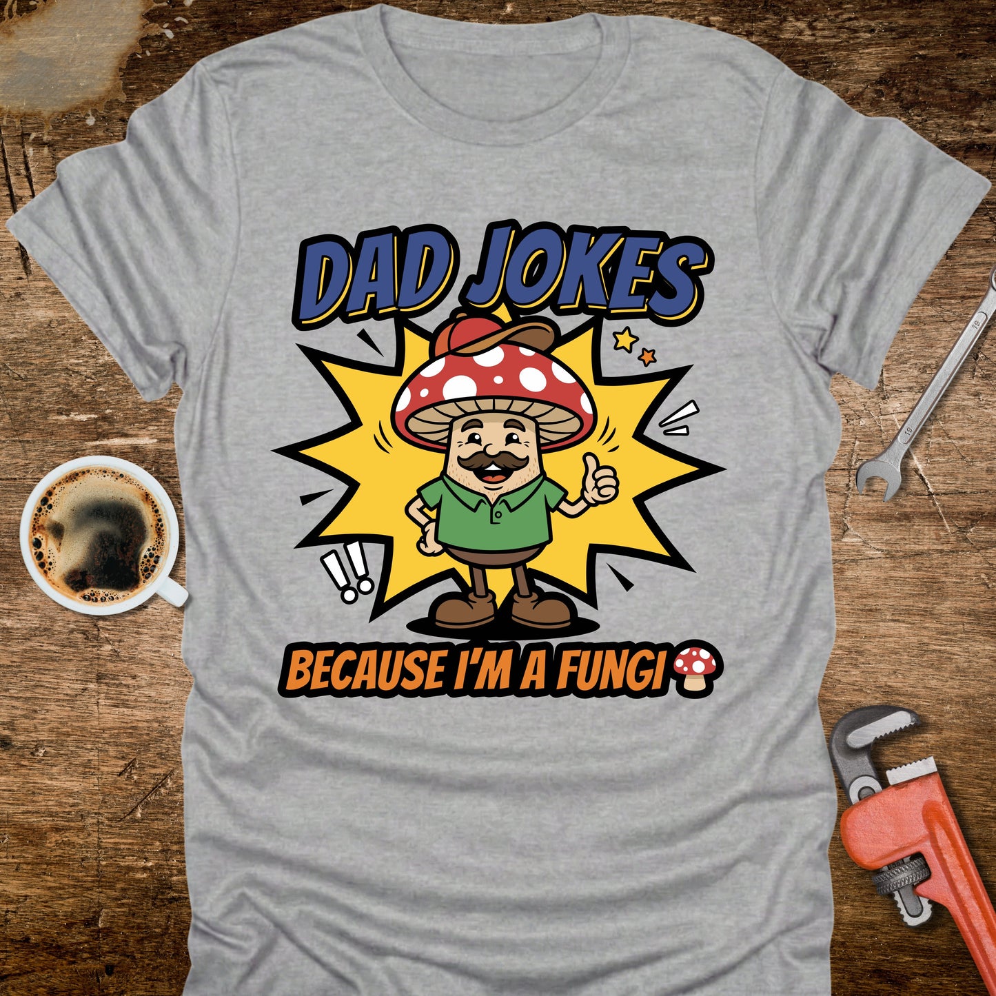 Gray t-shirt with a cartoon mushroom character and text on a wooden surface with coffee and tools.