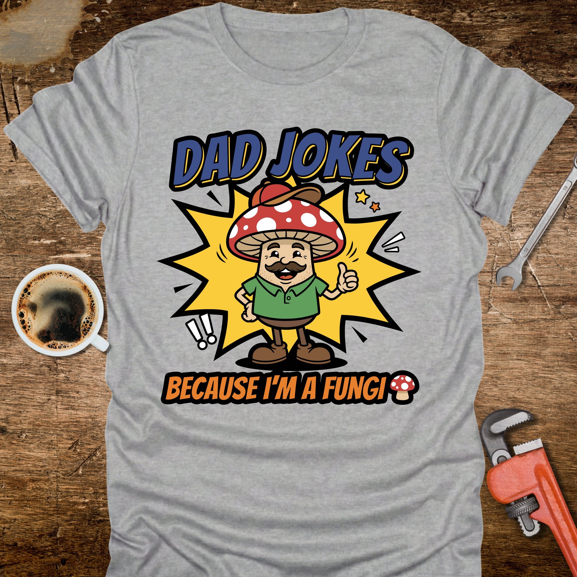 Gray t-shirt with a cartoon mushroom character and text on a wooden surface with coffee and tools.