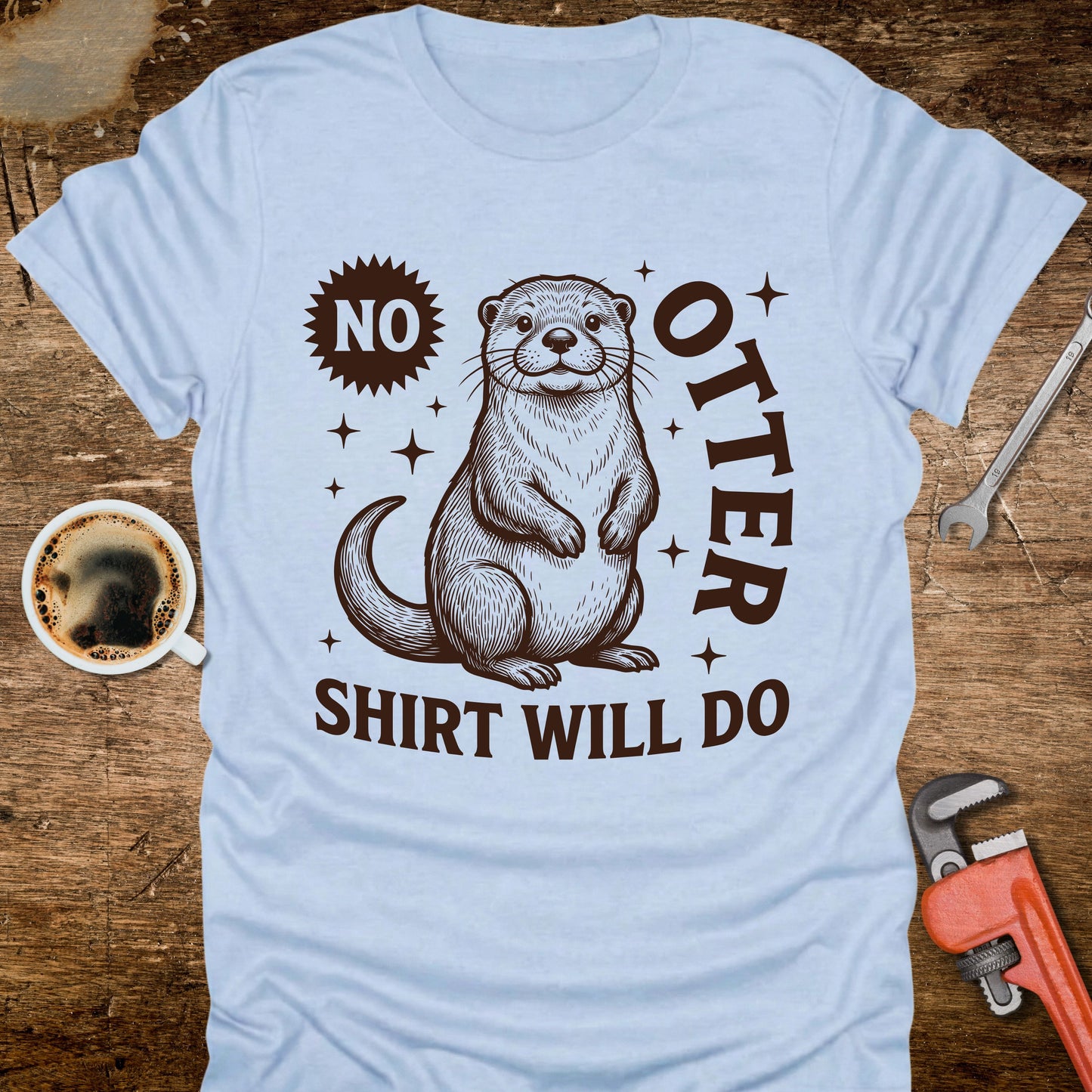 Light blue t-shirt with an otter graphic and text on a wooden surface with coffee and tools.