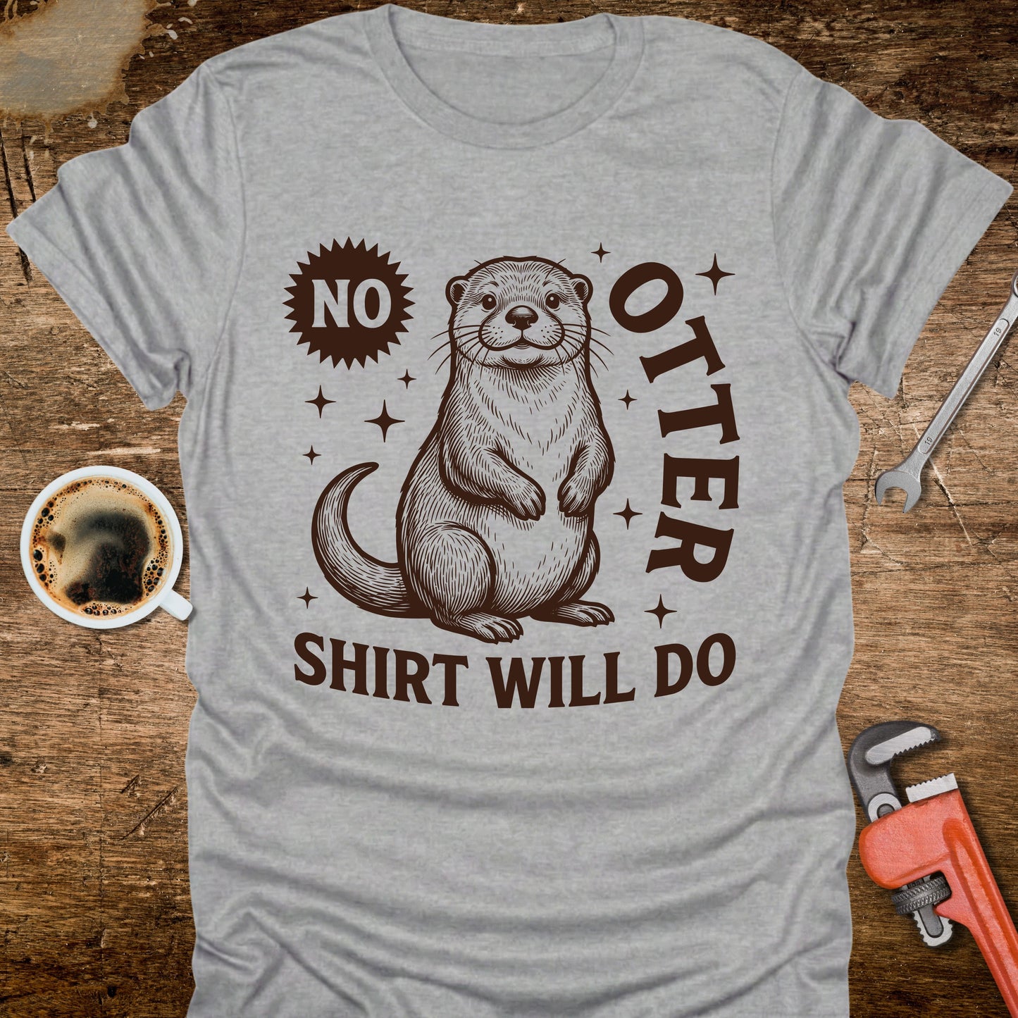 Gray t-shirt with an otter graphic and text on a wooden surface with coffee and tools.