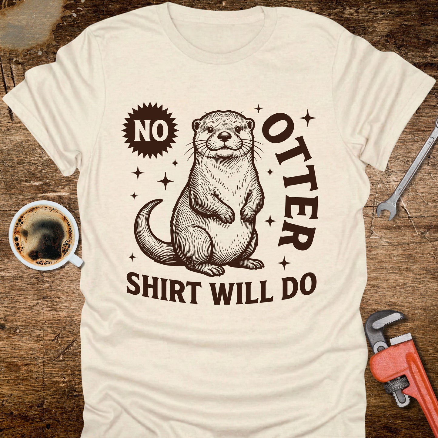Beige t-shirt with an otter graphic and text on a wooden surface with coffee and tools.