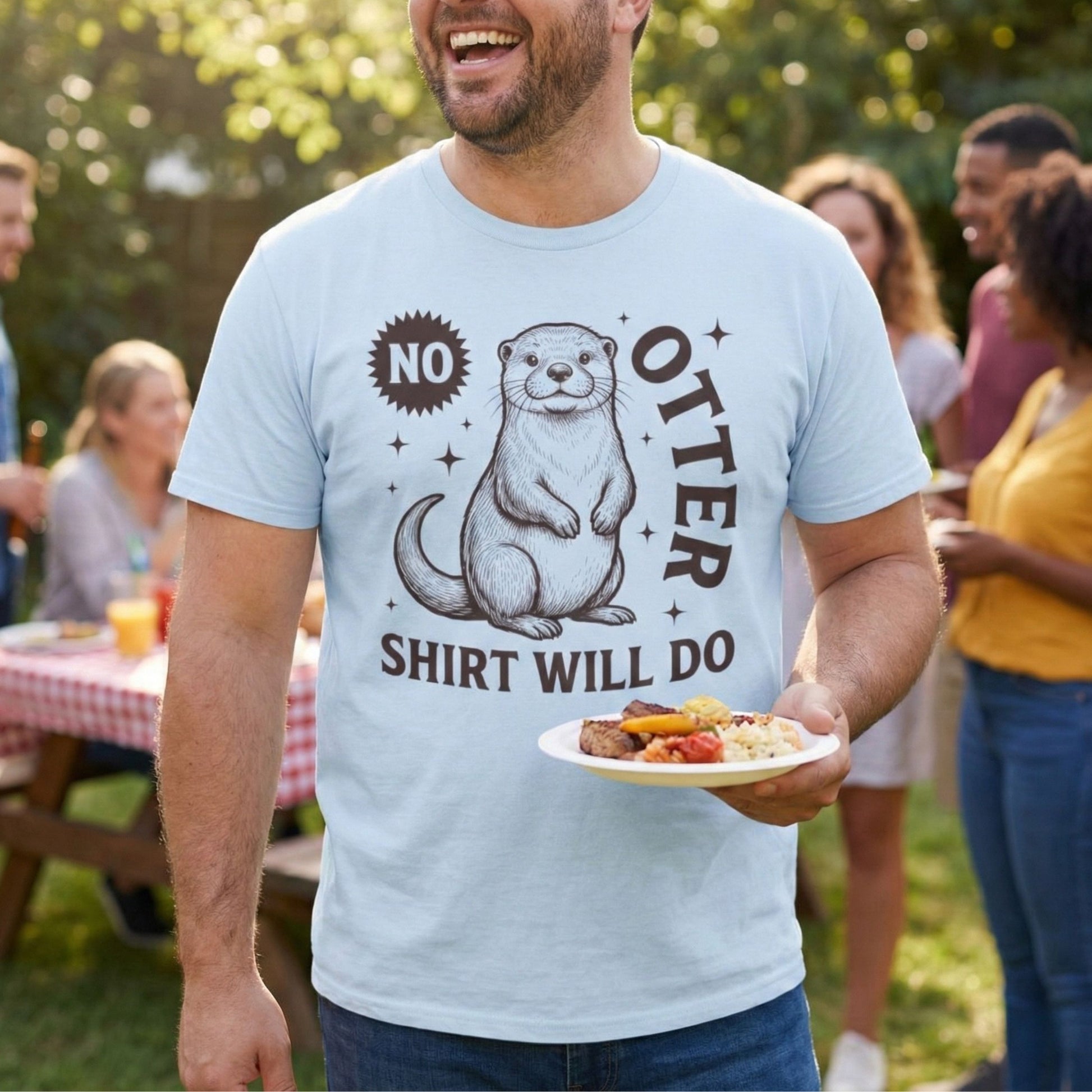Man wearing a t-shirt with an otter graphic and text, standing outdoors with people and a picnic table in the background.