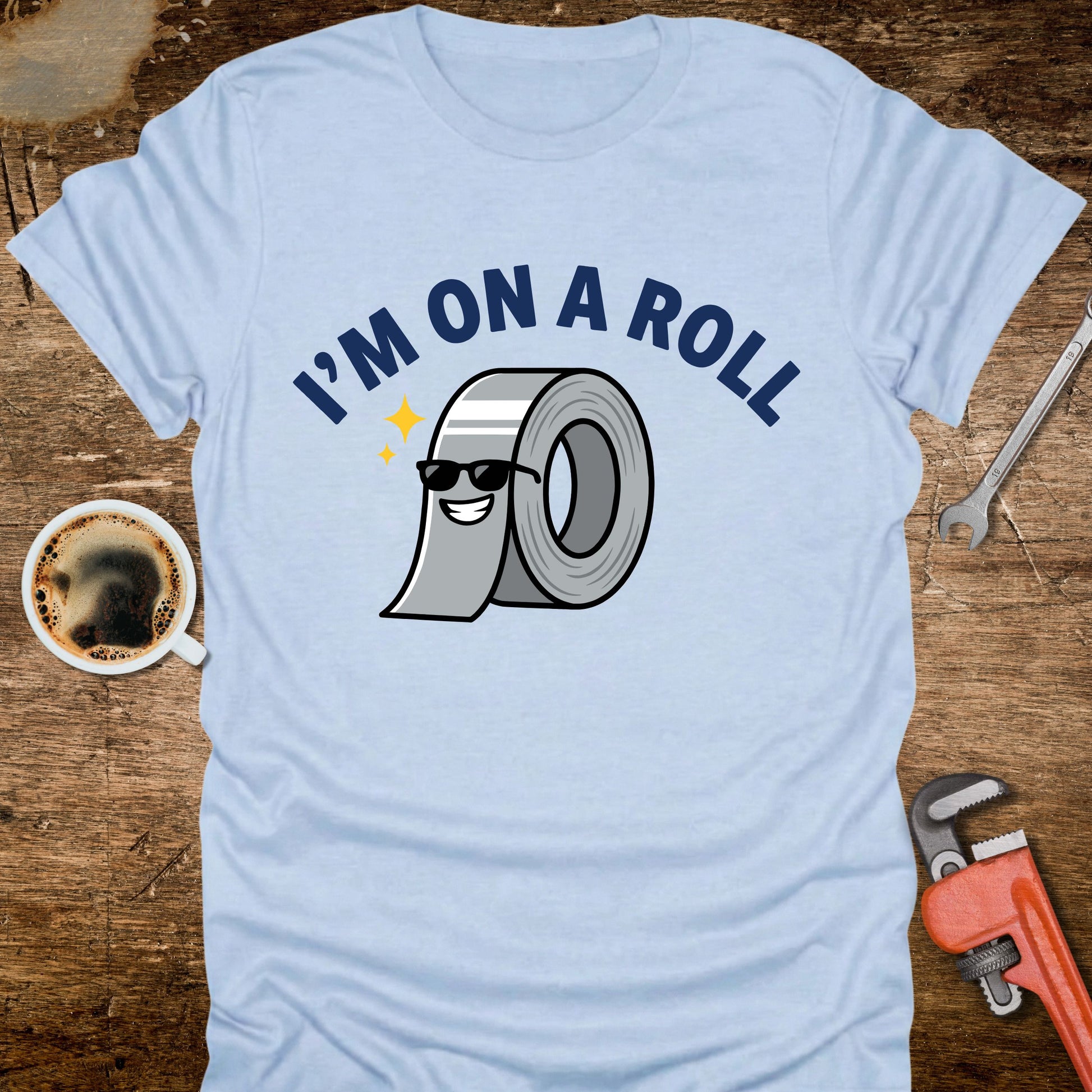 Light blue t-shirt with 'I'm on a Roll' graphic design featuring a roll of duct tape, placed on a wooden surface with tools and a coffee cup.