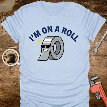 Light blue t-shirt with 'I'm on a Roll' graphic design featuring a roll of duct tape, placed on a wooden surface with tools and a coffee cup.