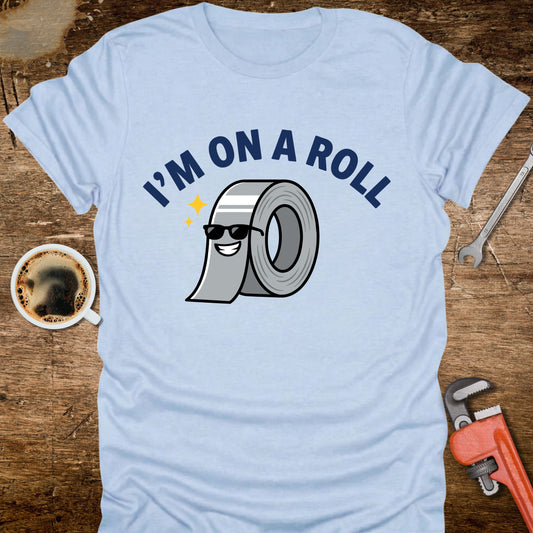 Light blue t-shirt with 'I'm on a Roll' graphic design featuring a roll of duct tape, placed on a wooden surface with tools and a coffee cup.