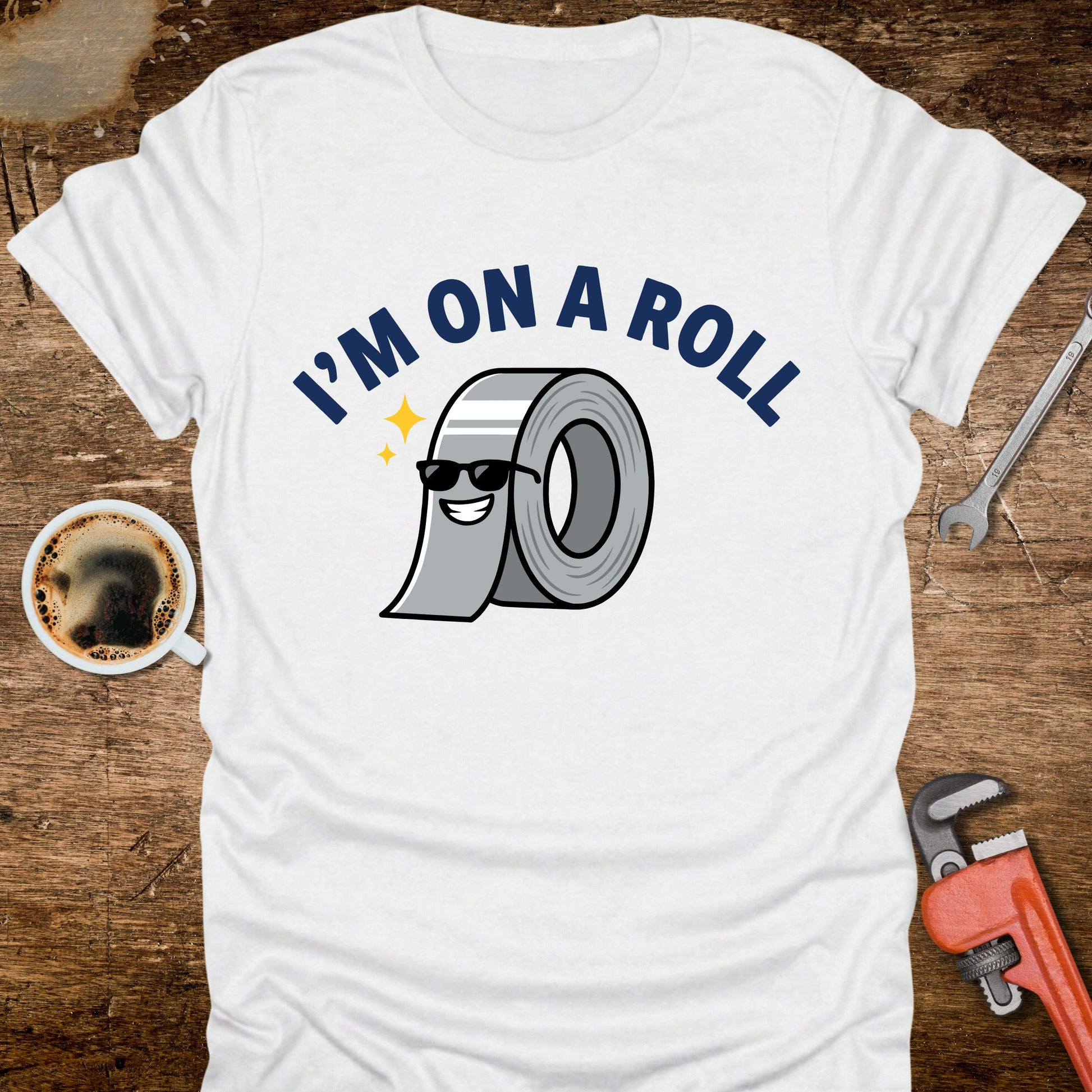 White t-shirt with 'I'm on a Roll' graphic design featuring a roll of duct tape, placed on a wooden surface with tools and a coffee cup.