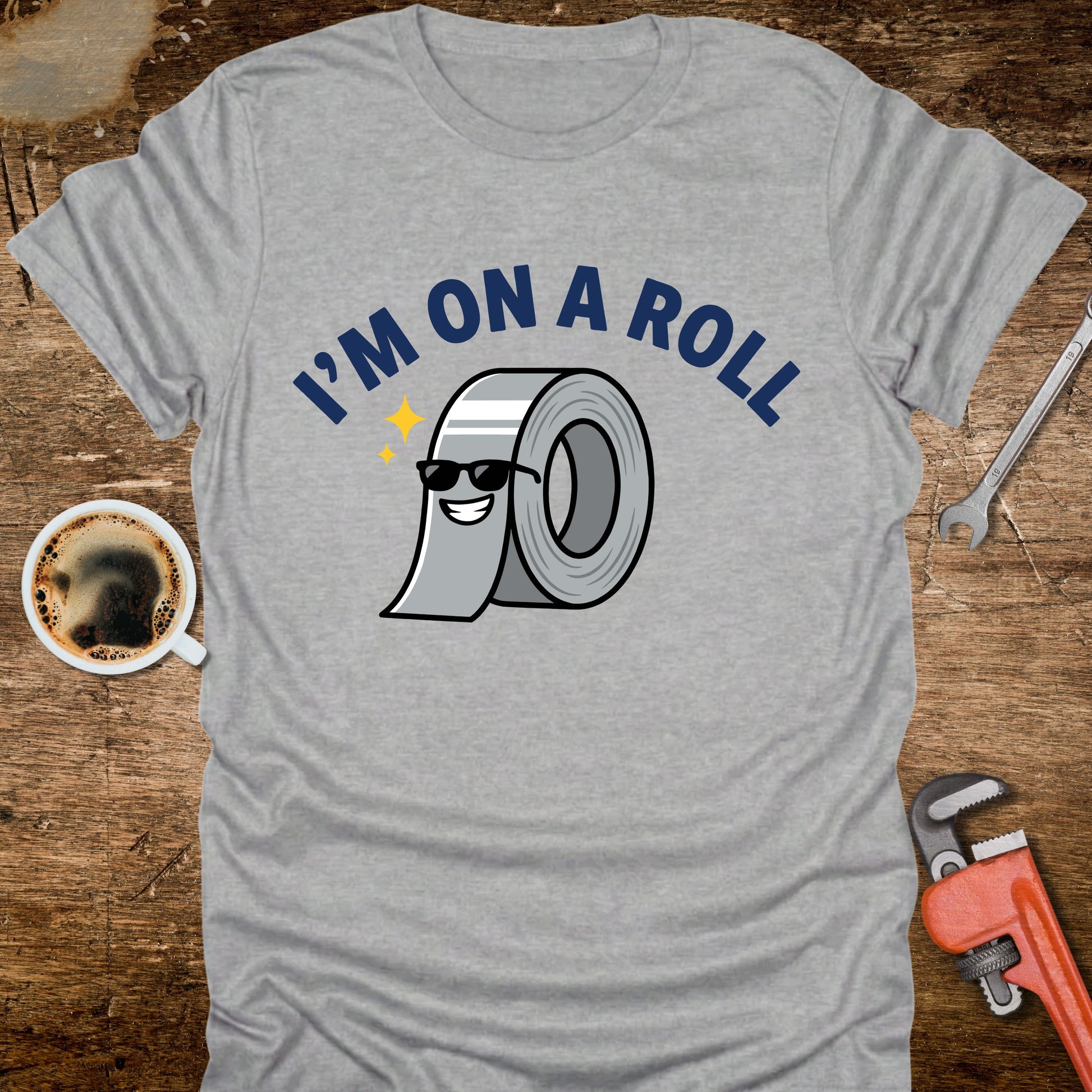 Gray t-shirt with 'I'm on a Roll' graphic design on a wooden surface with coffee and tools.