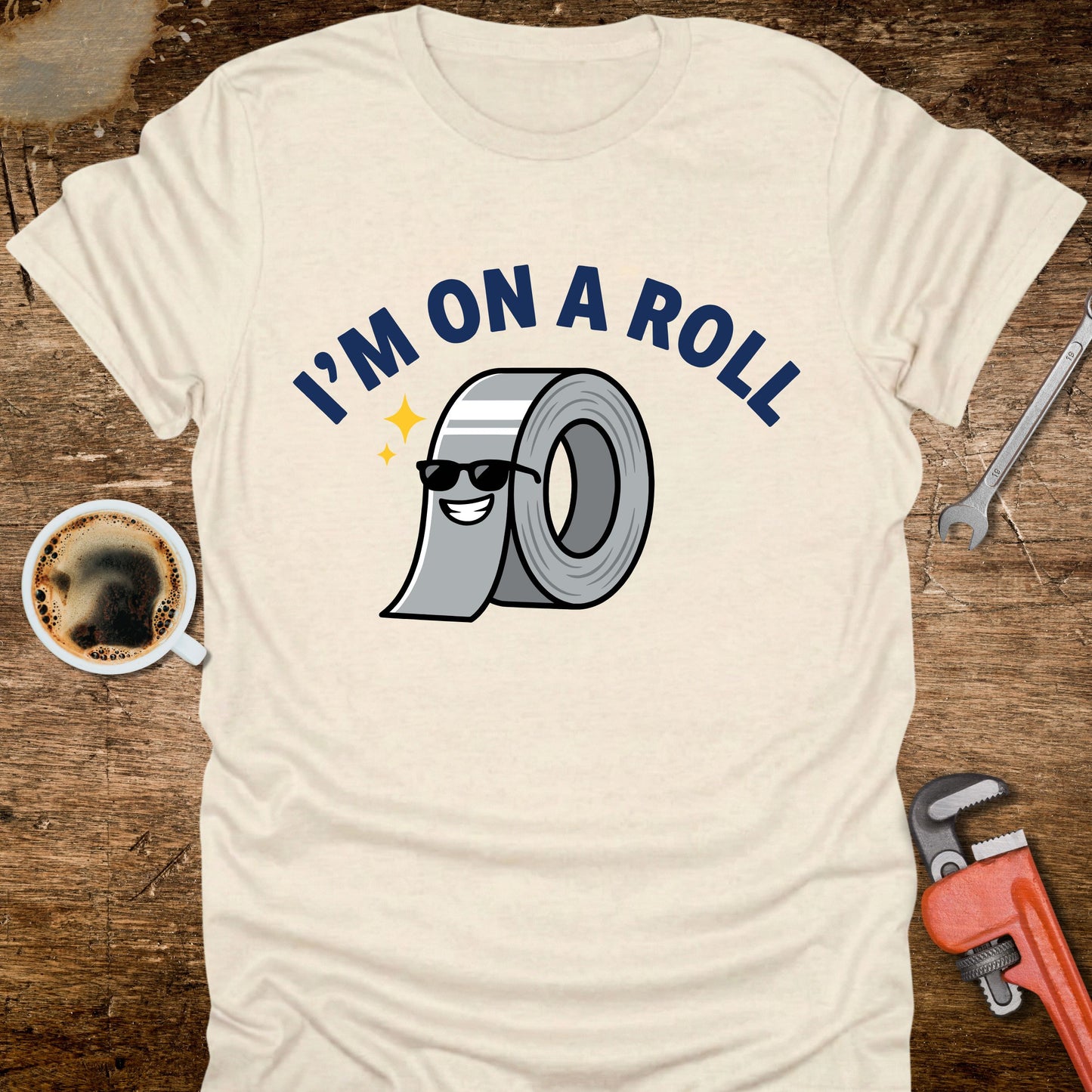 Beige t-shirt with 'I'm on a Roll' graphic design featuring duct tape, coffee cup, wrench, and pliers on a wooden surface.