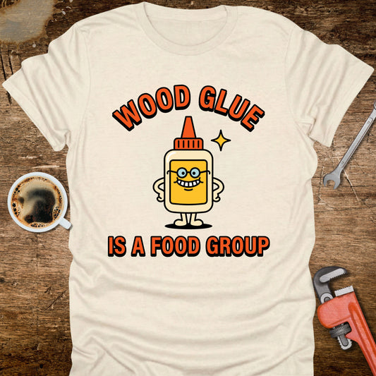 White t-shirt with a cartoon wood glue bottle graphic and text on a wooden surface with coffee and tools.