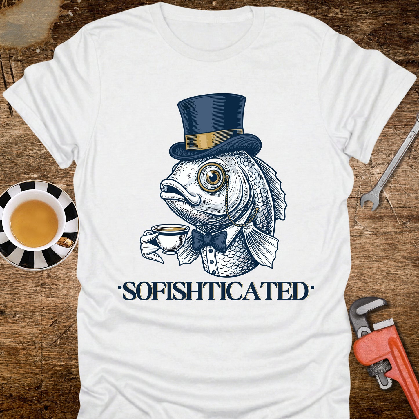 White t-shirt with a fish wearing a top hat and bow tie holding a cup, labeled 'Sofishticated', on a wooden surface with a cup and tools.