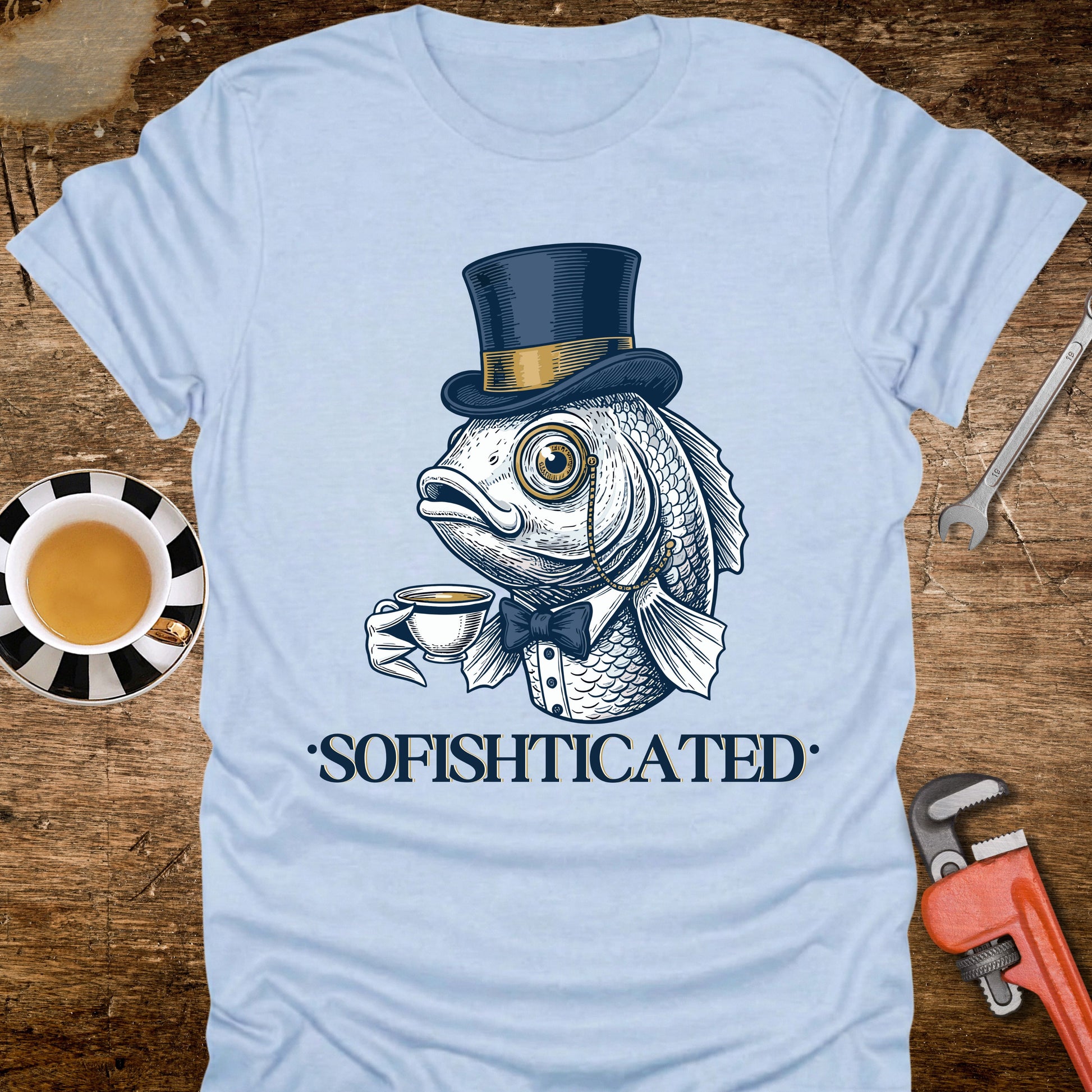 Light blue t-shirt with a fish wearing a top hat and bow tie holding a cup, labeled 'Sofishticated', on a wooden surface with a cup and tools.