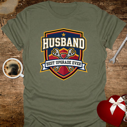 Green t-shirt with 'Husband Best Upgrade Ever' graphic on a wooden surface with coffee and heart-shaped gift box.