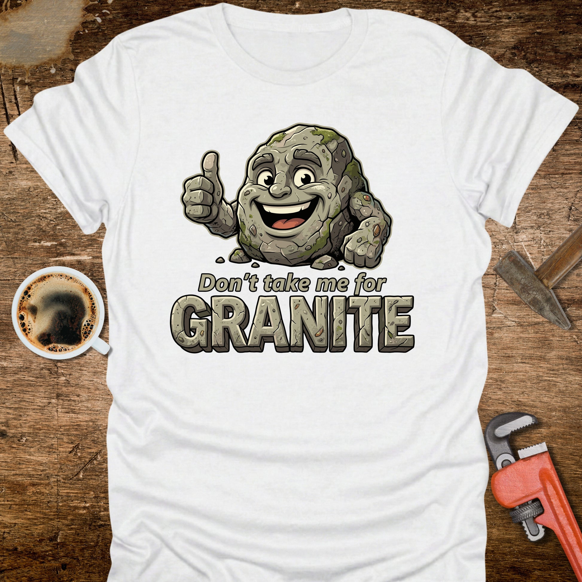 White t-shirt with a cartoon rock character and text 'Don't take me for Granite' on a wooden surface with a coffee cup and wrench.