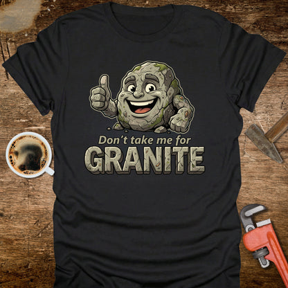 Black t-shirt with a cartoon stone character and text 'Don't take me for Granite' on a wooden surface with a coffee cup and wrench.