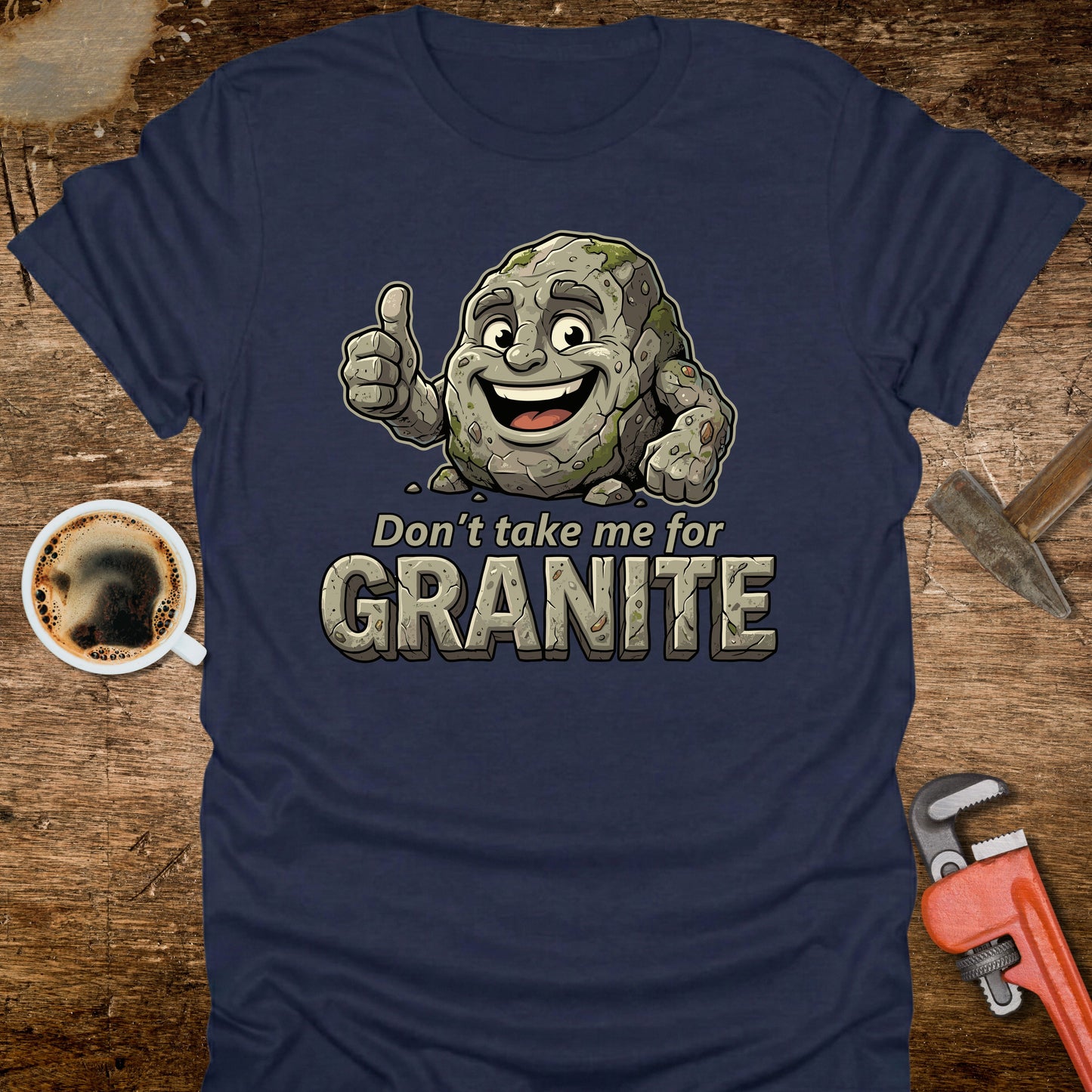Navy blue t-shirt with a cartoon stone character and text 'Don't take me for Granite' on a wooden surface with coffee and tools.