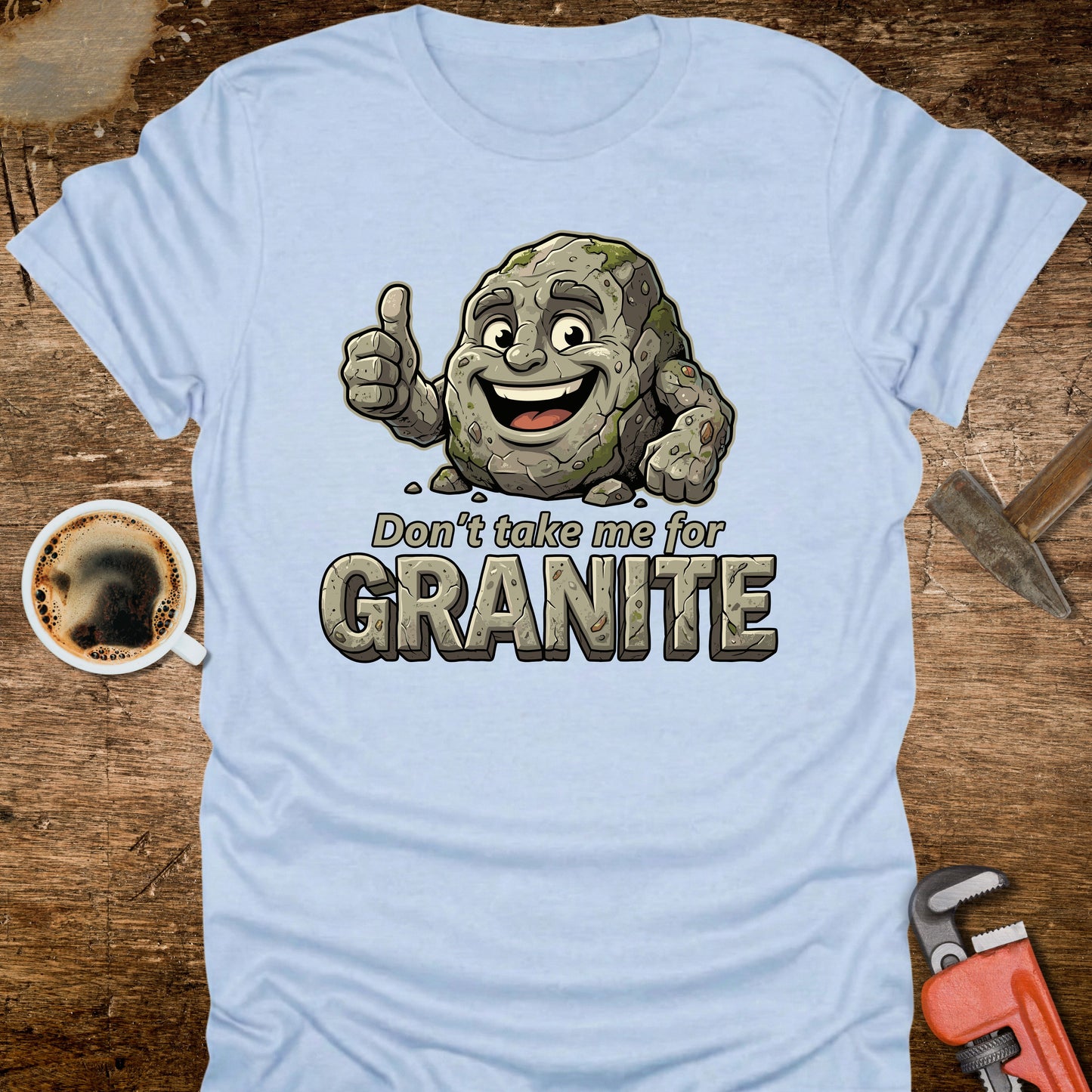 Light blue t-shirt with a cartoon rock character and text 'Don't take me for Granite' on a wooden surface with a coffee cup and wrench.