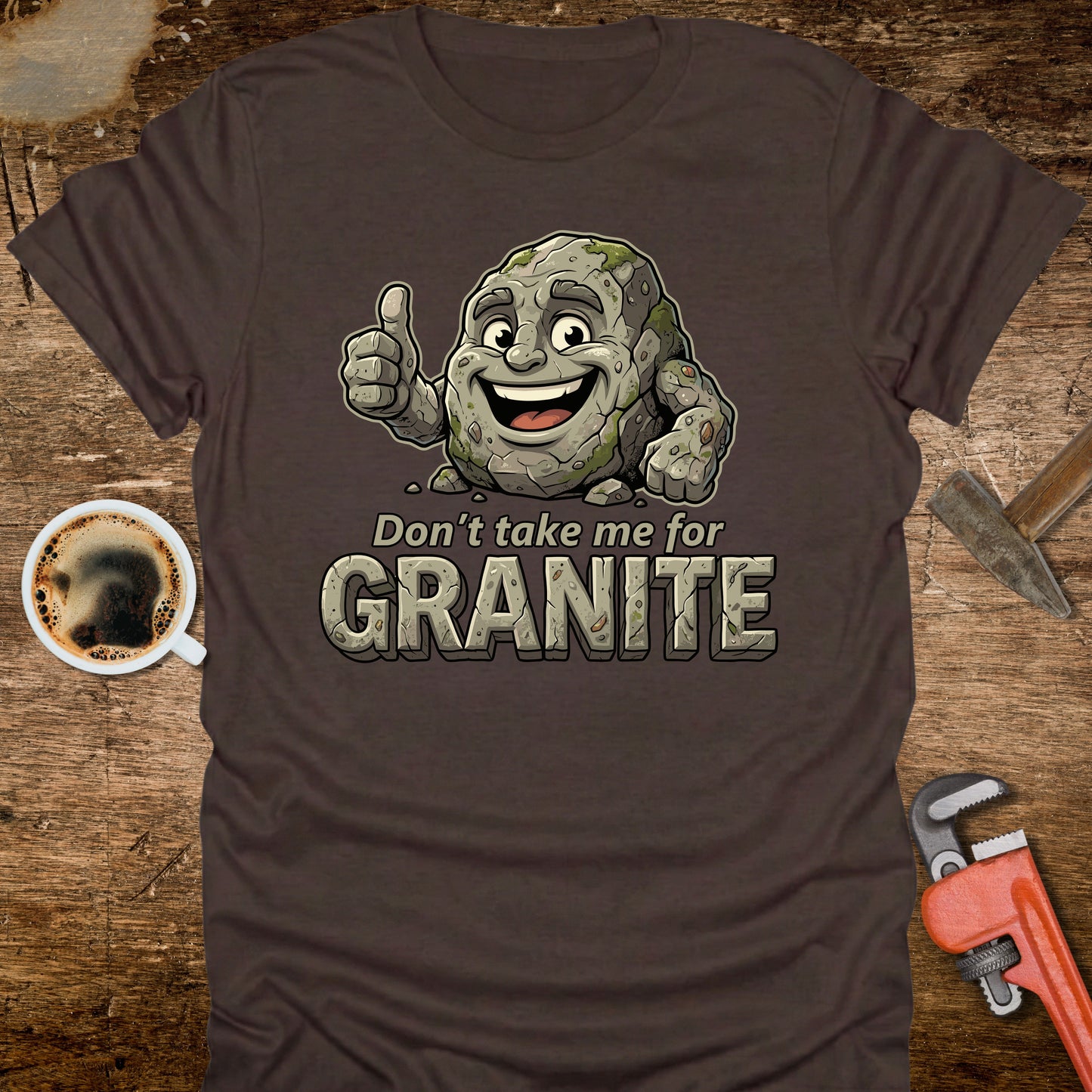 Brown t-shirt with a cartoon stone character and text 'Don't take me for Granite' on a wooden surface with a coffee cup and wrench.