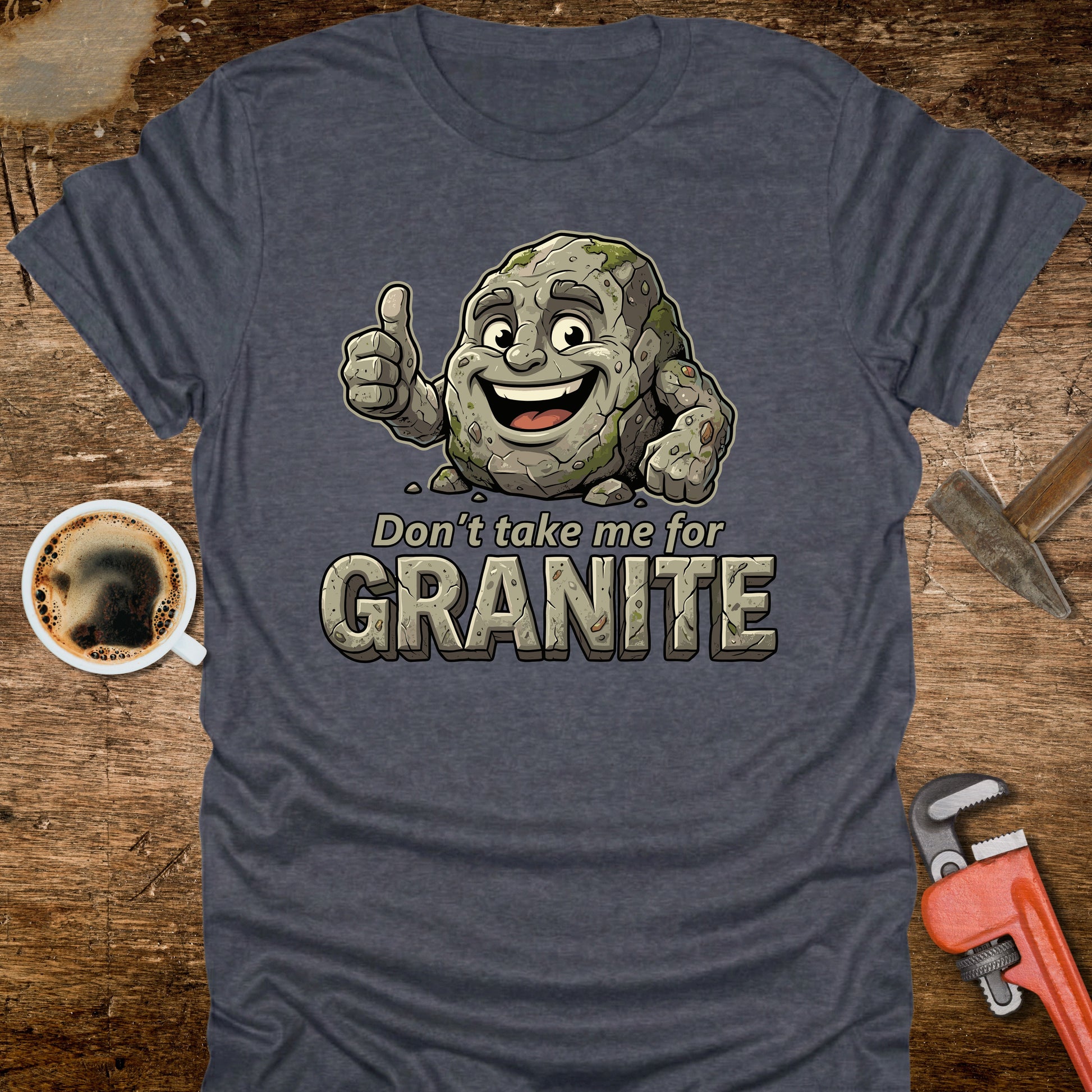 Blue t-shirt with cartoon granite character and text 'Don't take me for Granite' on a wooden surface with coffee and wrench.