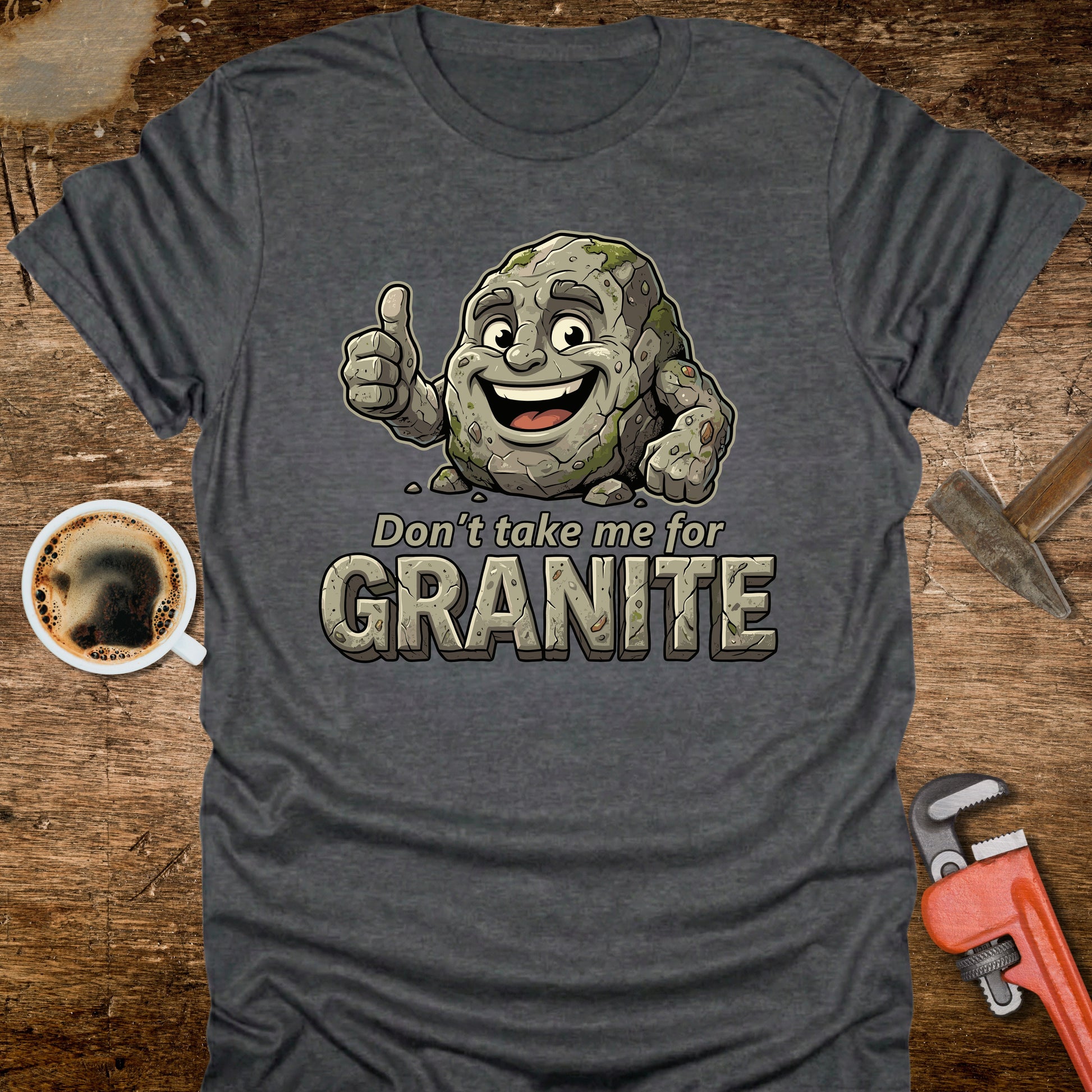 Gray t-shirt with cartoon character and 'Don't take me for Granite' text on a wooden surface with coffee and wrench.