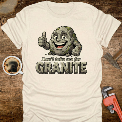 White t-shirt with a cartoon rock character and text 'Don't take me for Granite' on a wooden surface with coffee and a wrench.