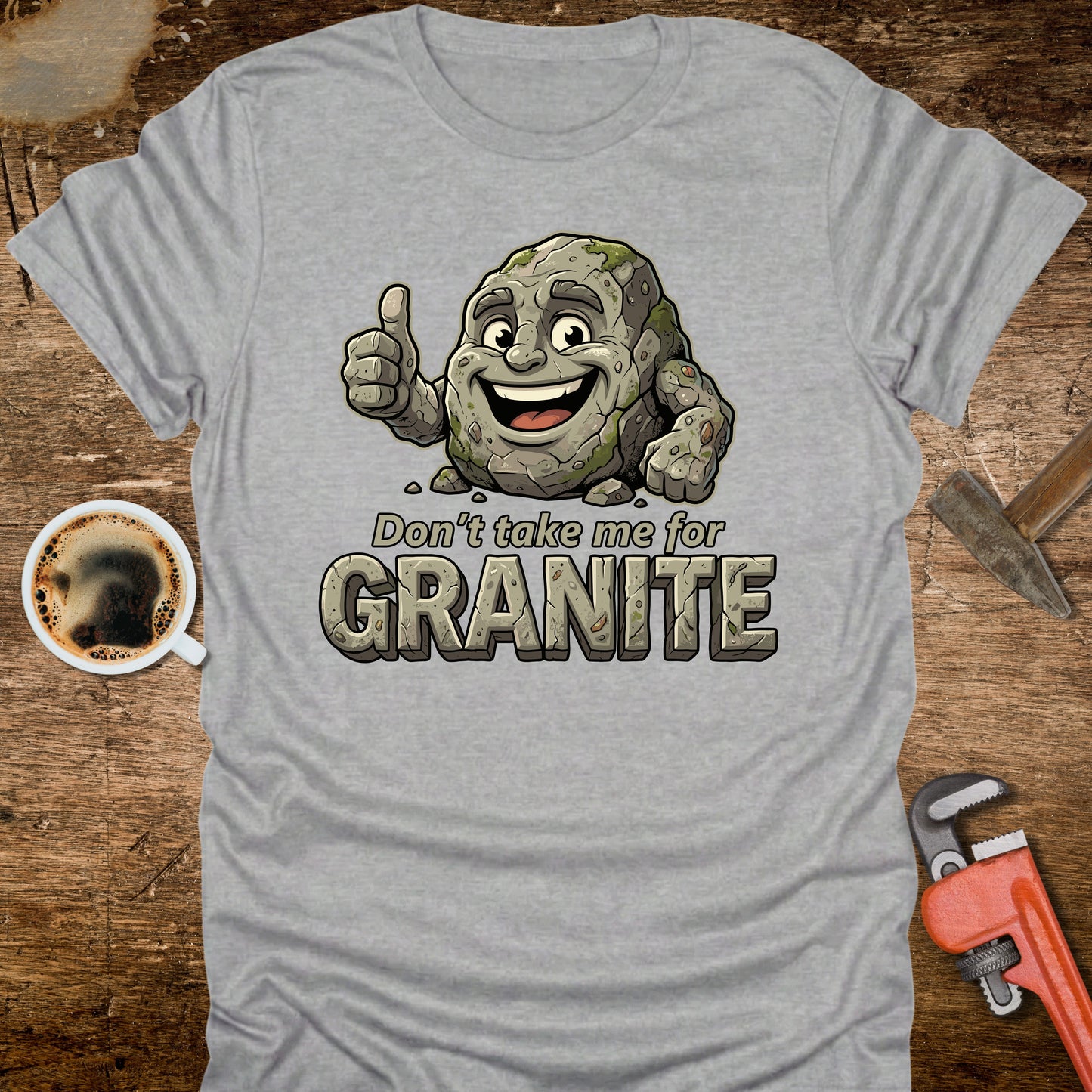 Gray t-shirt with a cartoon rock character and text 'Don't take me for Granite' on a wooden surface with a coffee cup and wrench.