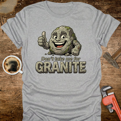 Gray t-shirt with a cartoon rock character and text 'Don't take me for Granite' on a wooden surface with a coffee cup and wrench.