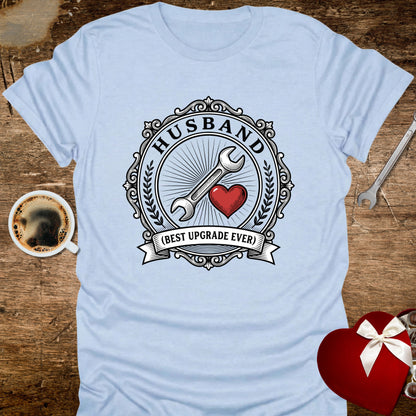 Light blue t-shirt with 'Husband (Best Upgrade Ever)' design on a wooden surface with coffee and gift.