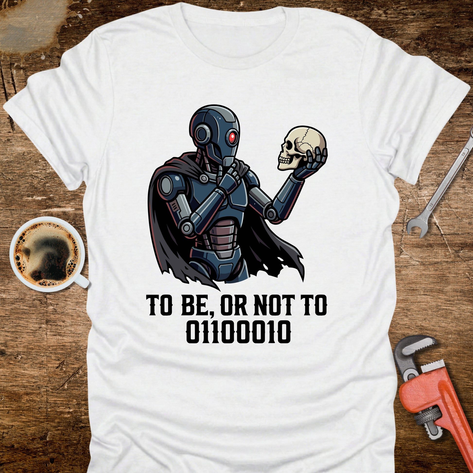White t-shirt with robot and skull design on a wooden surface with tools and coffee.