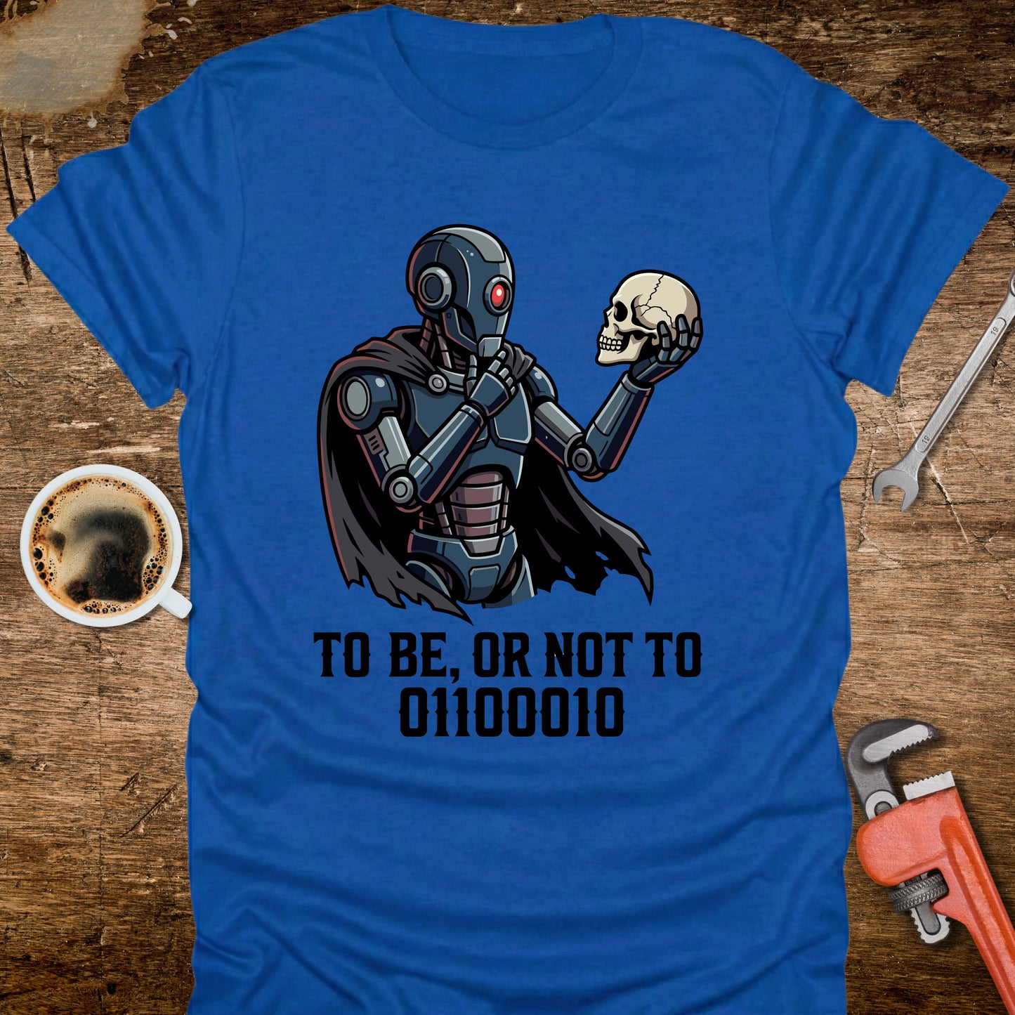 Blue t-shirt with robot holding a skull design and text on a wooden surface with tools and coffee.