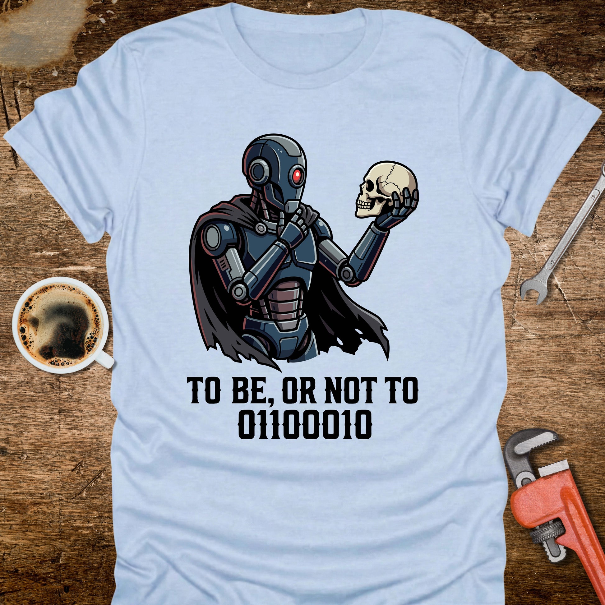 Light blue t-shirt with a robot holding a skull and text on a wooden surface with coffee and tools.
