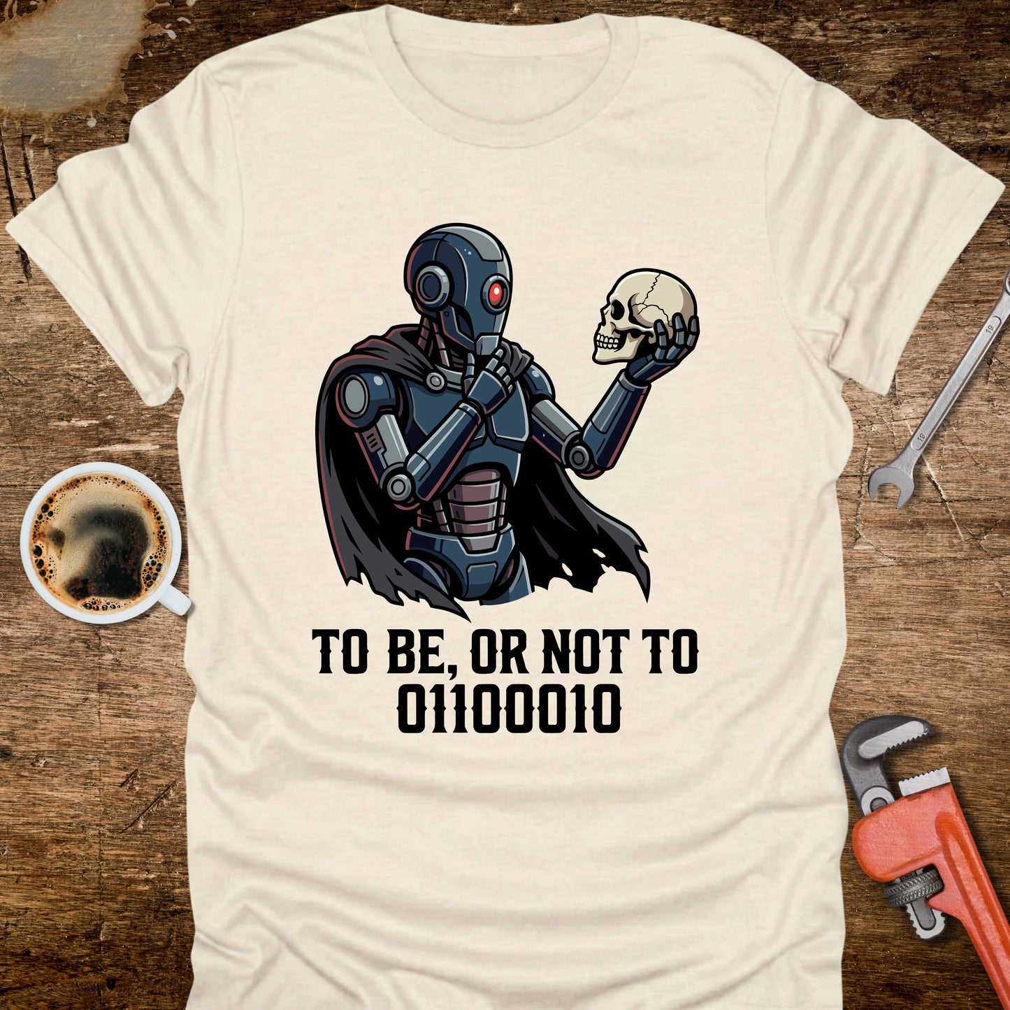 T-shirt with robot holding a skull design and text on a wooden surface with coffee and tools.