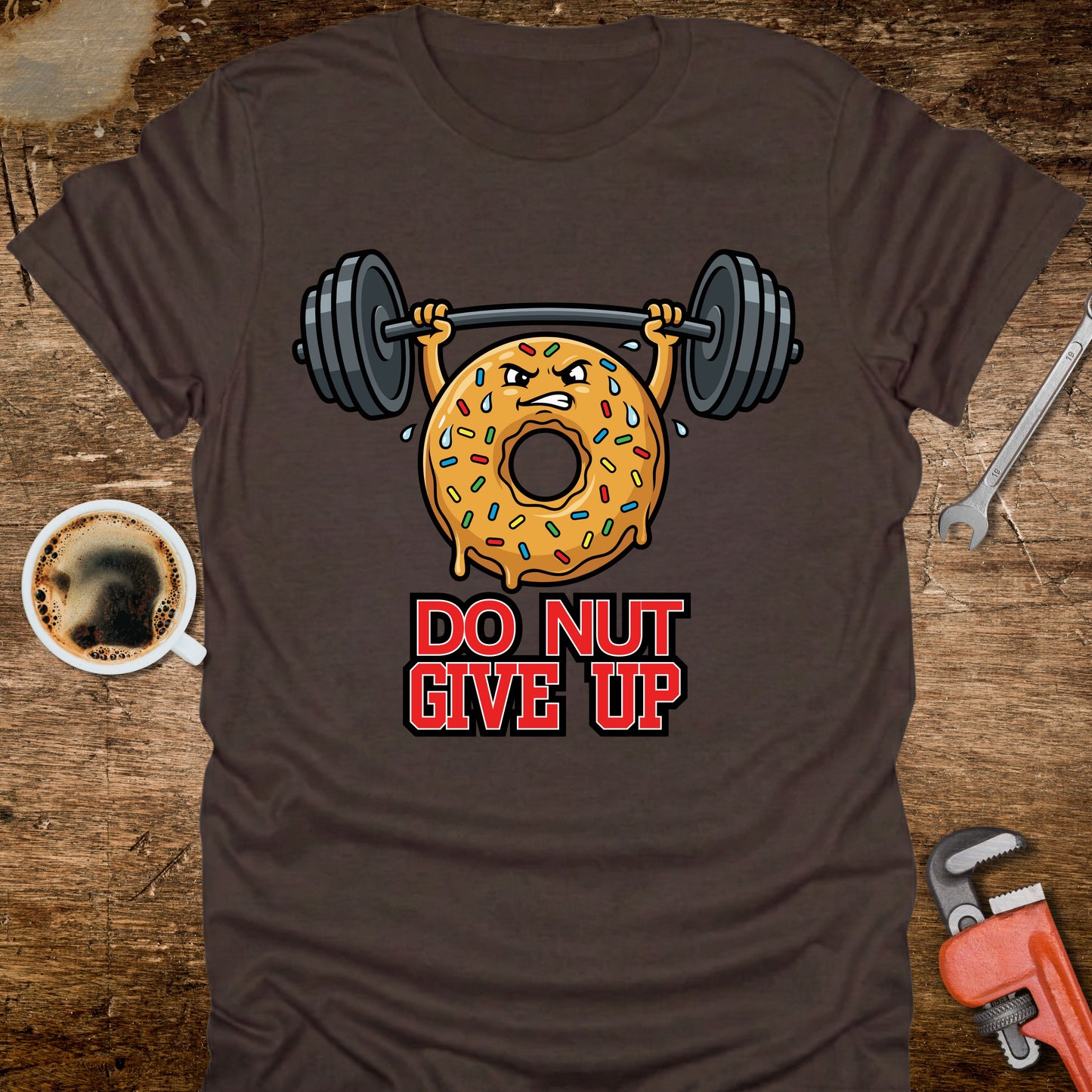 Brown t-shirt with a donut lifting weights graphic and 'Do Nut Give Up' text on a wooden surface.