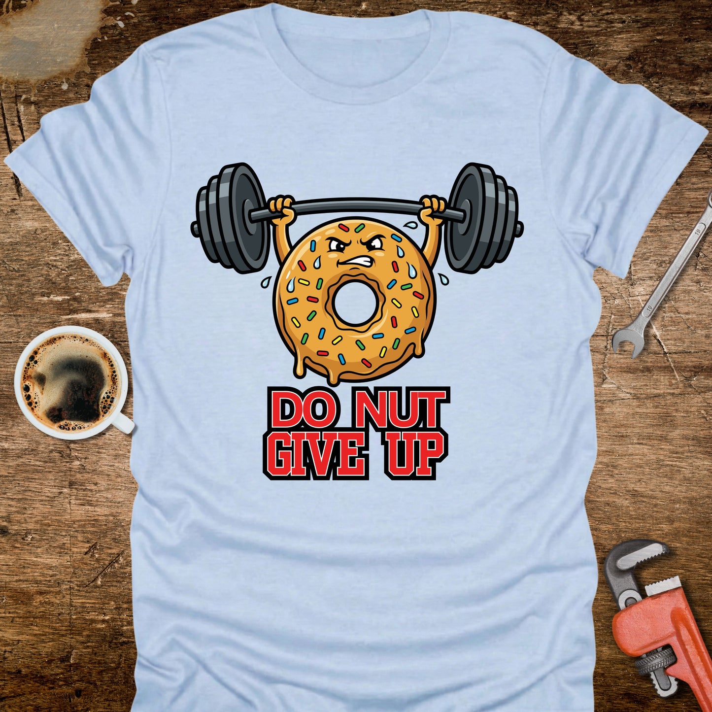 Light blue t-shirt with a donut lifting weights and 'Do Nut Give Up' text on a wooden surface with coffee and tools.
