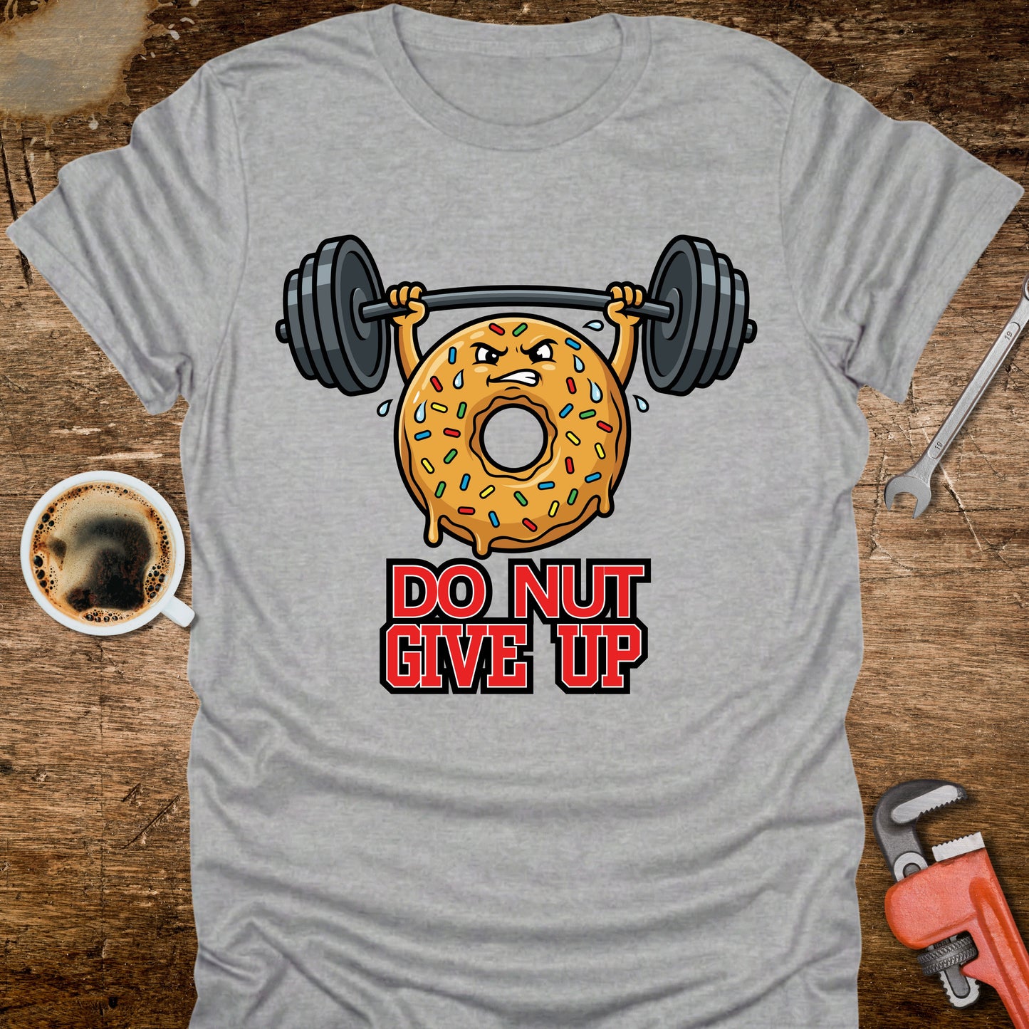 Gray t-shirt with a donut lifting weights and 'Do Nut Give Up' text on a wooden surface with coffee and tools.