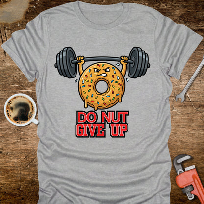 Gray t-shirt with a donut lifting weights and 'Do Nut Give Up' text on a wooden surface with coffee and tools.