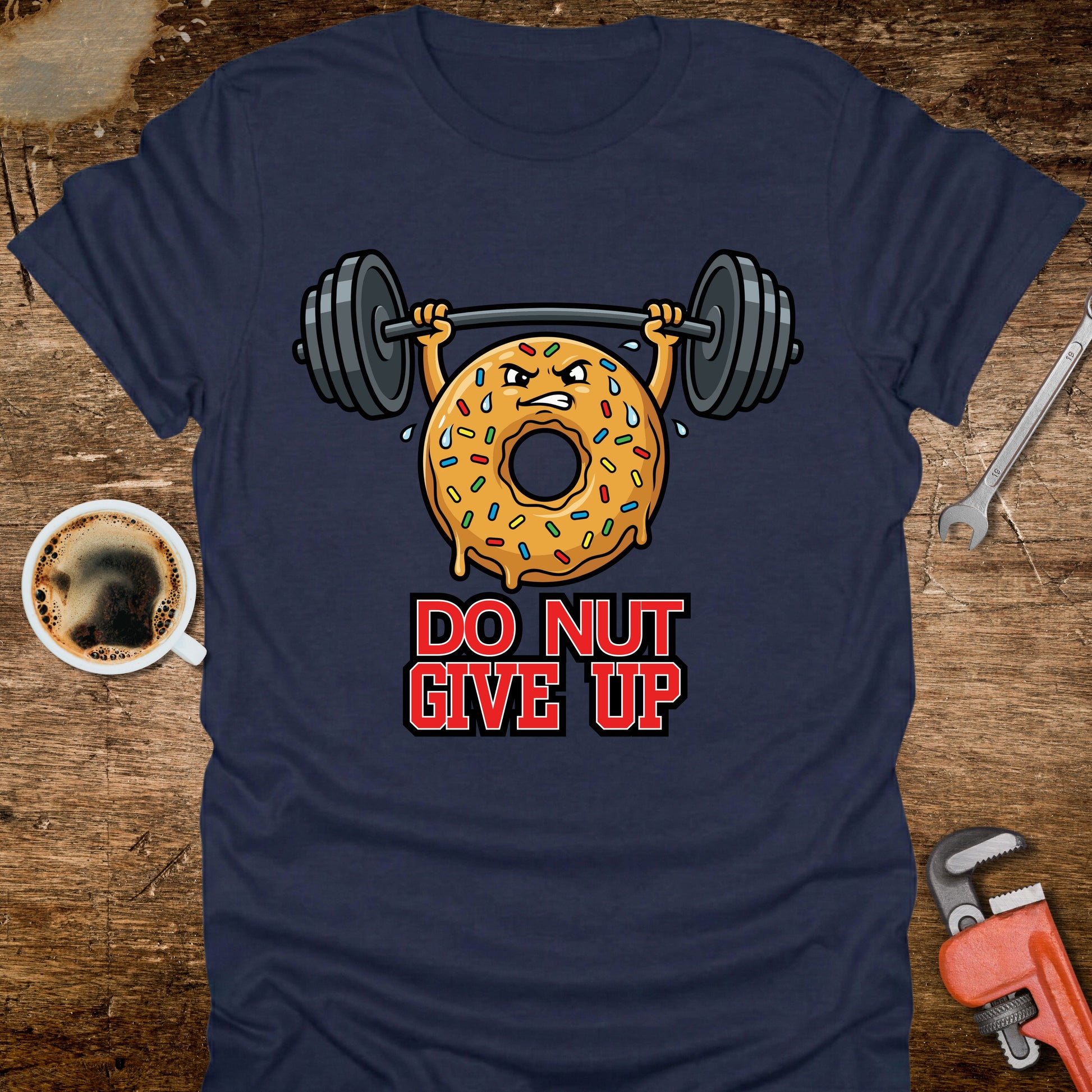 Navy blue t-shirt with a donut lifting weights graphic and 'Do Nut Give Up' text on a wooden surface with coffee and tools.