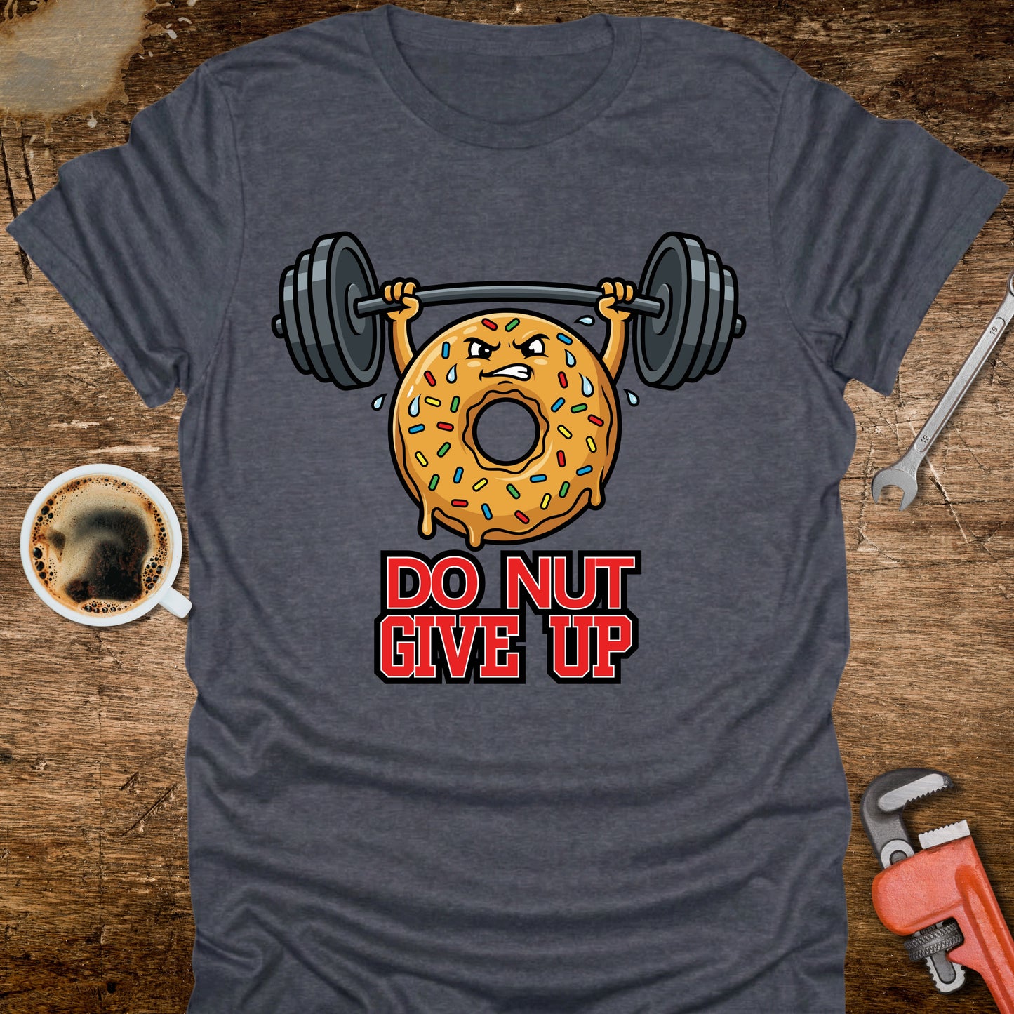 Blue t-shirt with a donut lifting weights graphic and 'Do Nut Give Up' text on a wooden surface with coffee and tools.