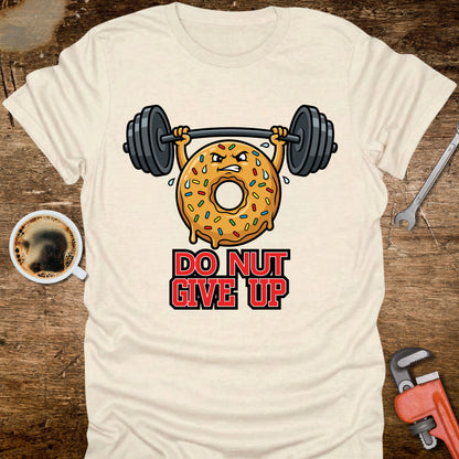Beige t-shirt with a donut lifting weights graphic and 'Do Nut Give Up' text on a wooden surface with coffee and tools.
