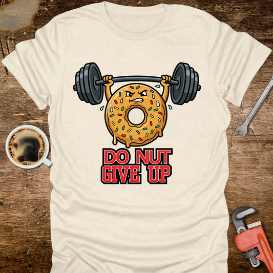 Beige t-shirt with a donut lifting weights graphic and 'Do Nut Give Up' text on a wooden surface with coffee and tools.