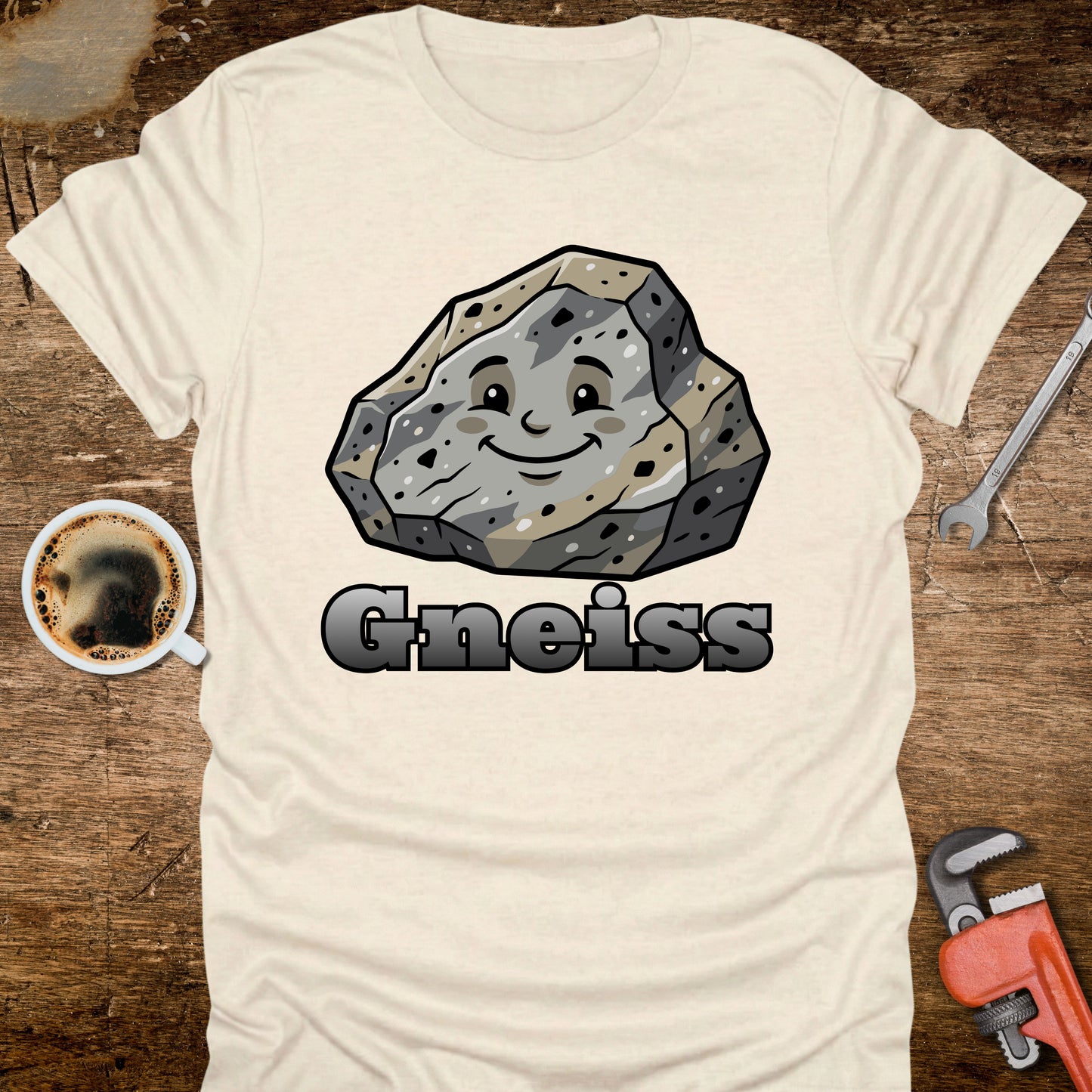 White t-shirt with a cartoon gneiss rock graphic and the word 'Gneiss' on a wooden surface with tools and coffee.