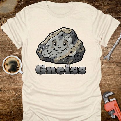White t-shirt with a cartoon gneiss rock graphic and the word 'Gneiss' on a wooden surface with tools and coffee.